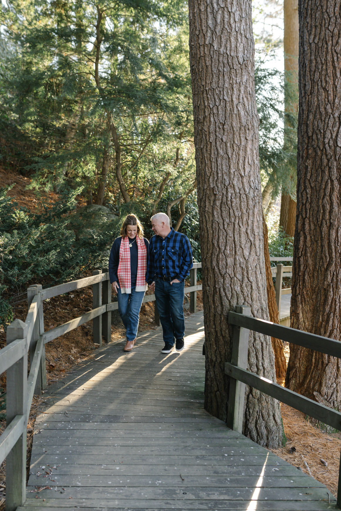 A man and woman walking together on a wooden boardwalk through a forested area, holding hands and smiling.
