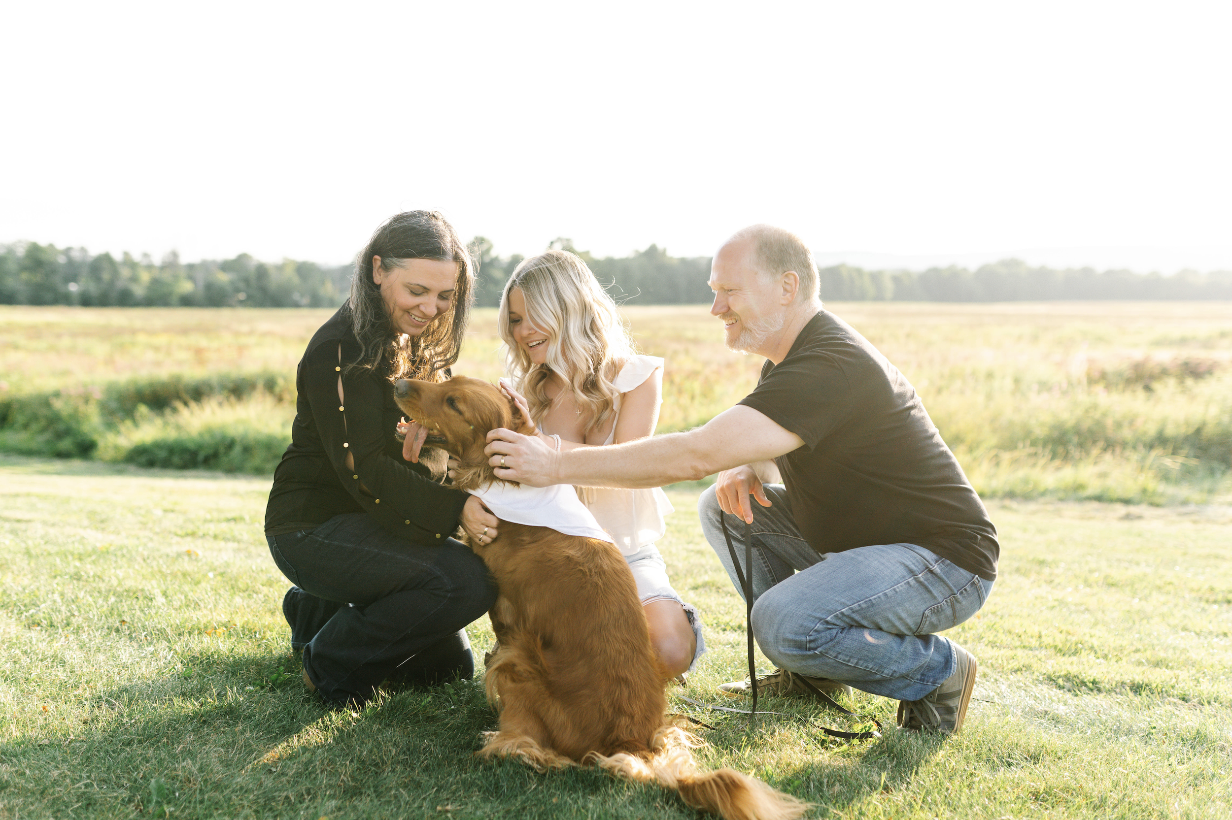 A family of three with a dog in a grassy field on a sunny day, smiling and playing with the dog.