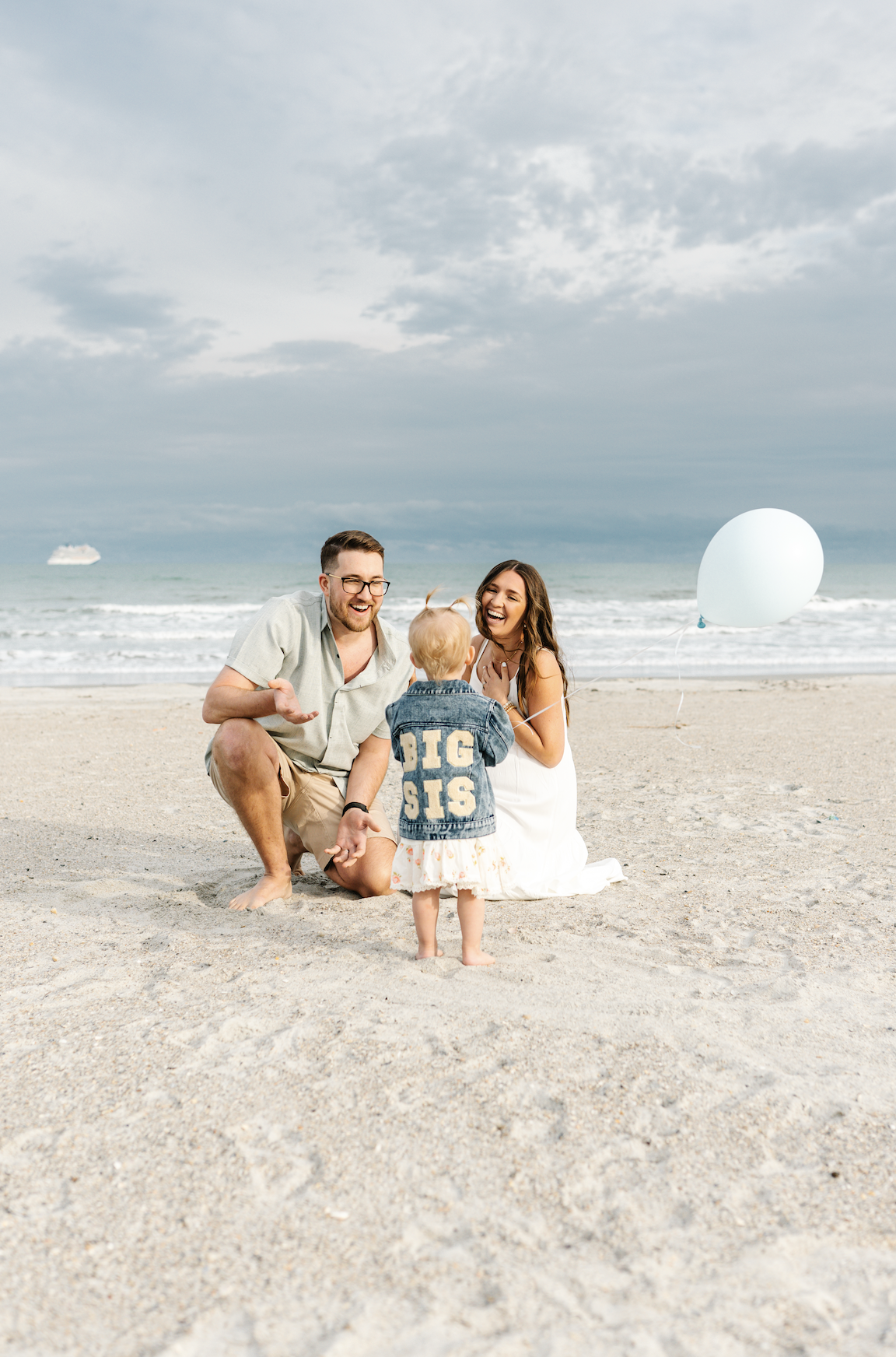 A family of three with a young girl in a denim jacket on a sandy beach. The father and mother are kneeling and smiling at their child, who is standing between them. The child has blonde hair and is wearing a dress and a jacket with 'BIG SIS' on it. T