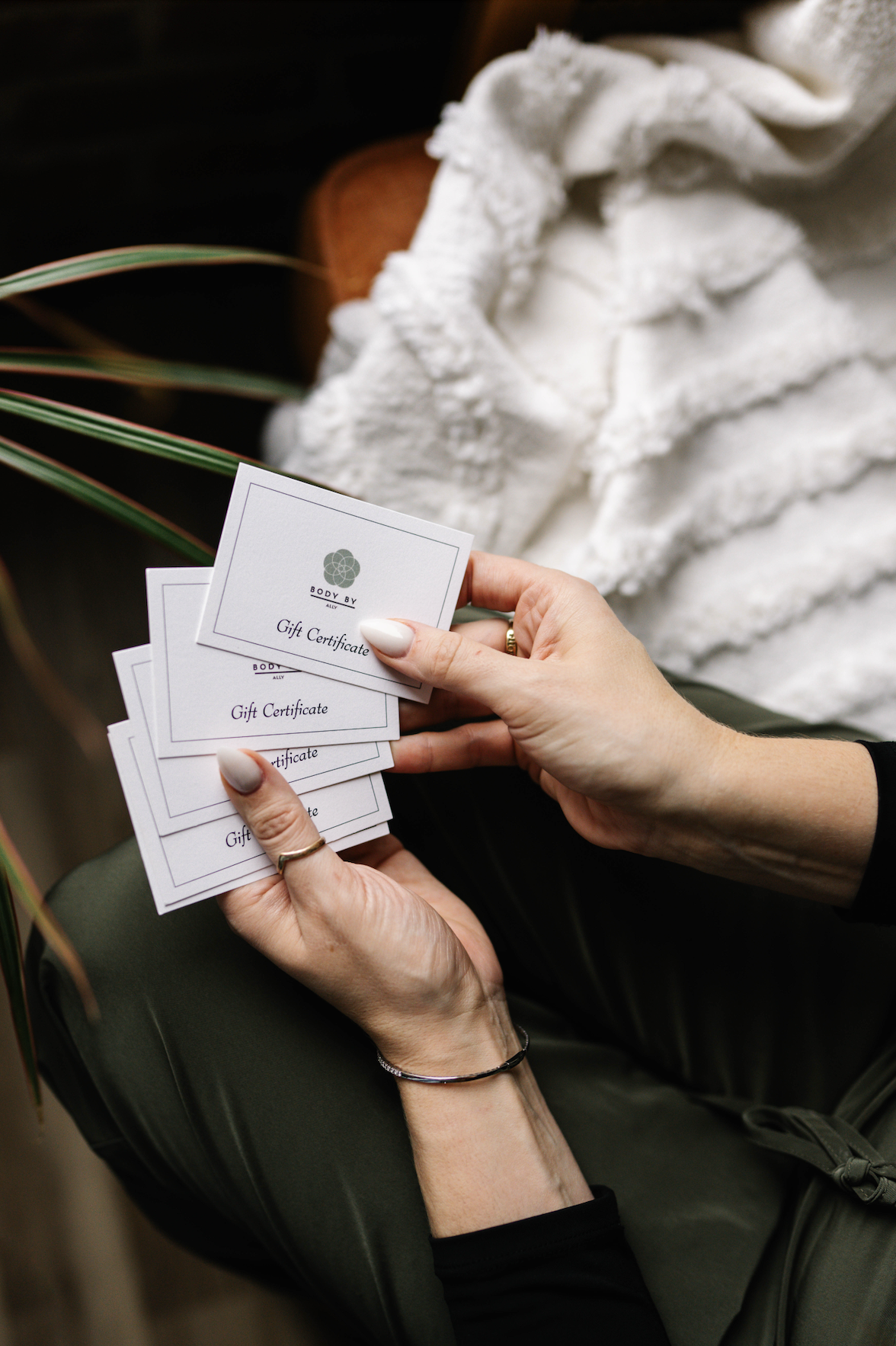 Hands holding multiple gift certificates for Body By Ally, with a white fluffy blanket in the background.