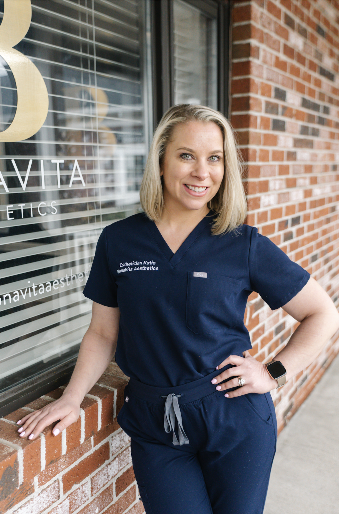 A woman in navy scrubs with 'Esthetician Katie BonaVita Aesthetics' embroidered on her shirt standing outside a brick building. She has blonde hair, is smiling, and is wearing a smartwatch.