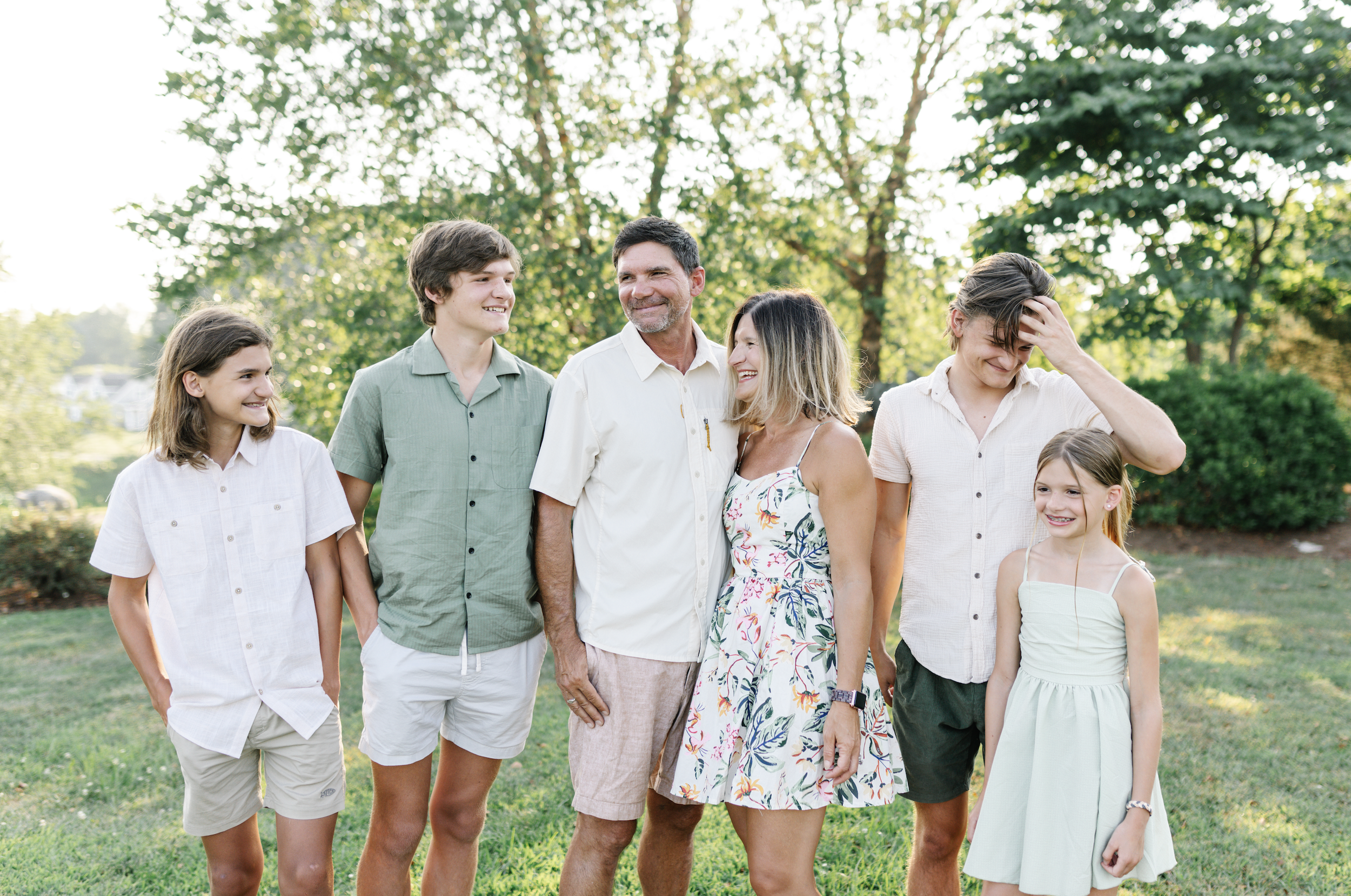 Family of seven standing outdoors on lush green grass with trees in the background, smiling and interacting during daytime.