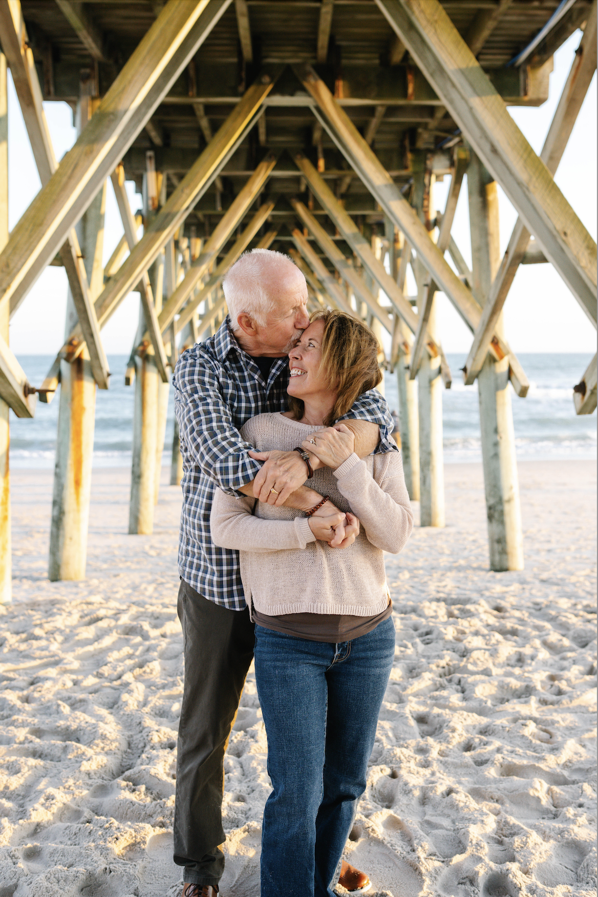 An elderly man kissing a woman on the forehead under a wooden pier at the beach, smiling and embracing. The ocean is visible in the background.