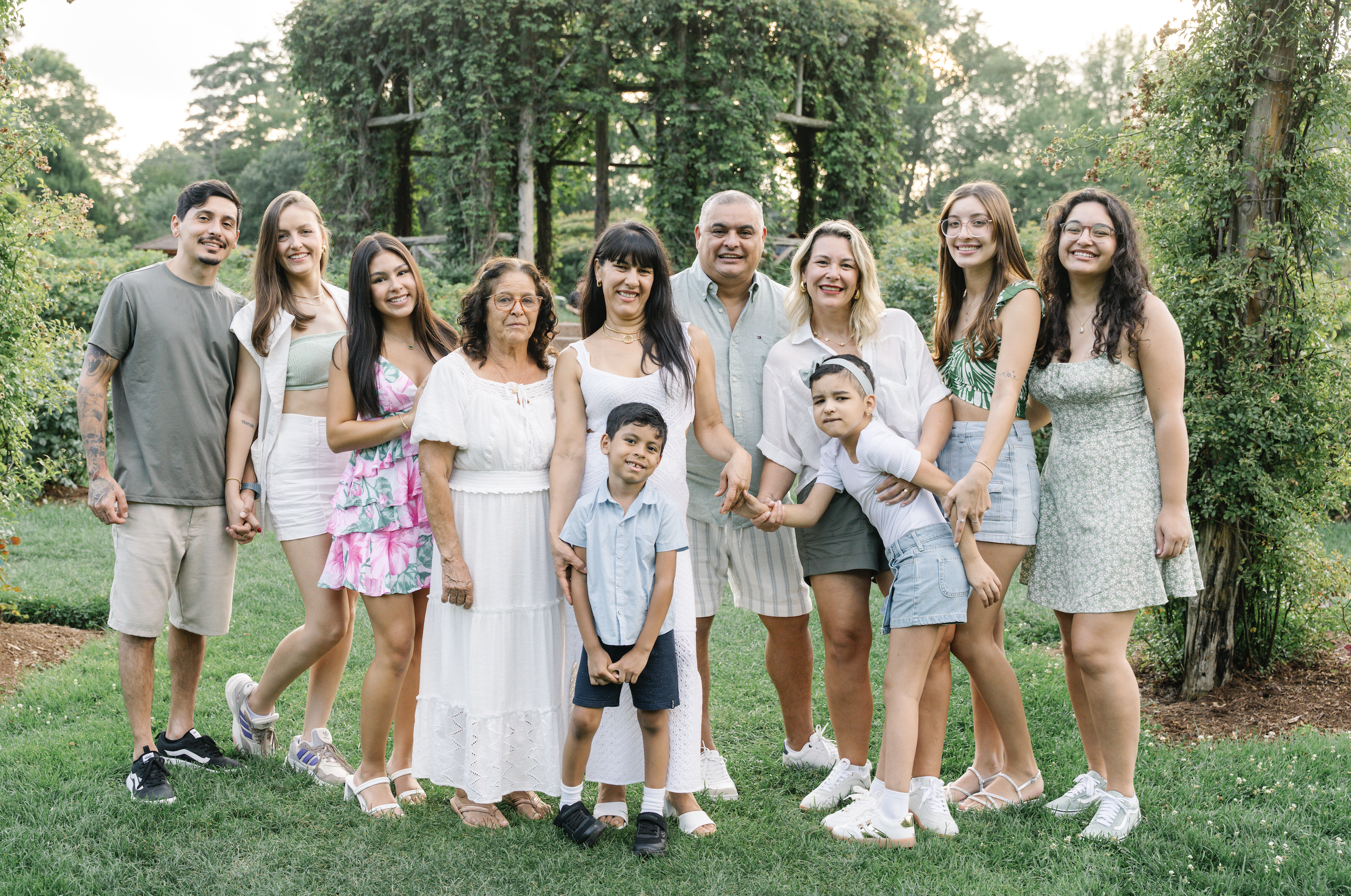 A diverse group of 12 people, including children and adults, standing outdoors on a grassy area with trees in the background, posing for a photo and smiling.