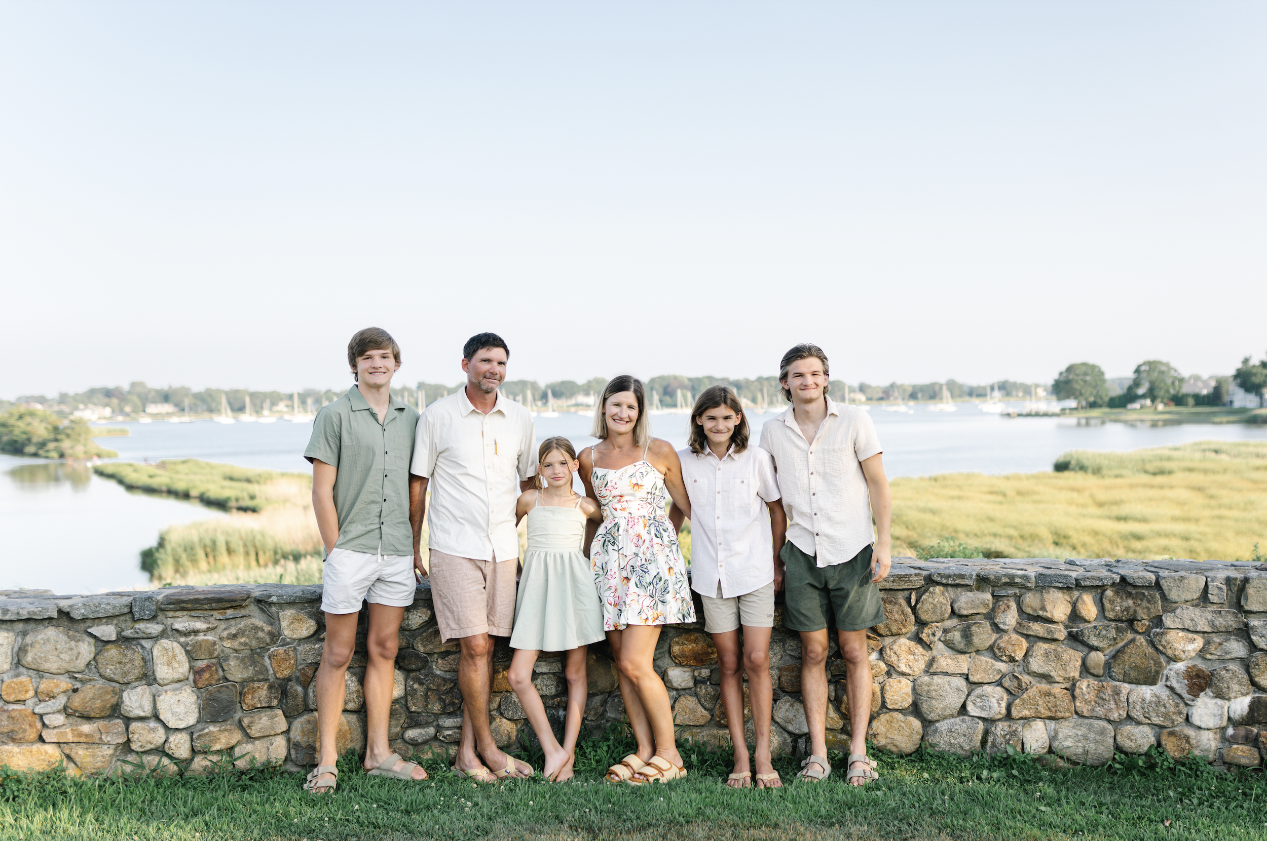 A family of seven standing behind a stone wall at a lake, smiling, with sailboats and trees in the background, during daytime.