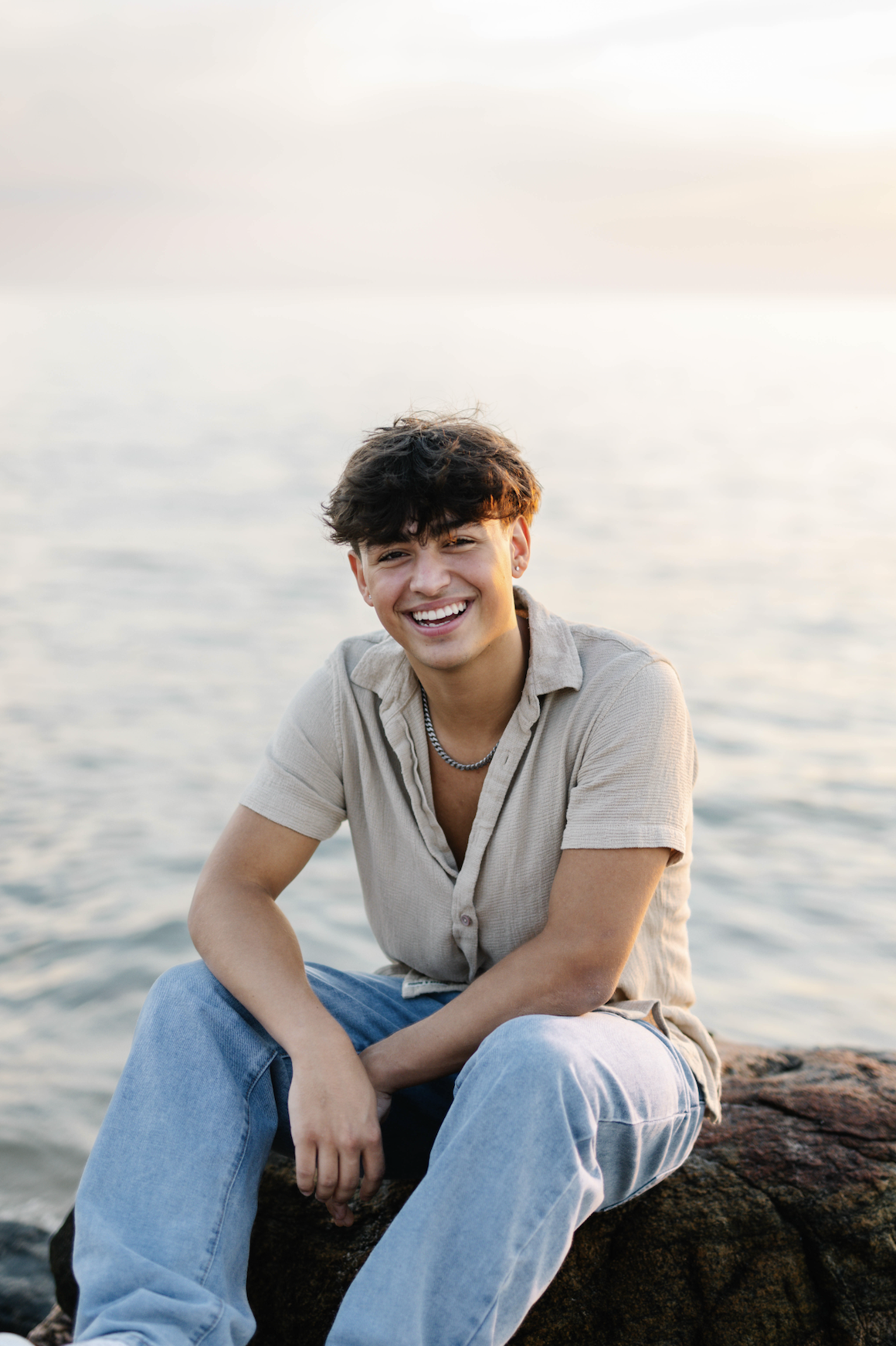 A young man with dark, curly hair, wearing a beige short-sleeve shirt and light blue jeans, is sitting on a rock by a body of water, smiling at the camera during sunset.