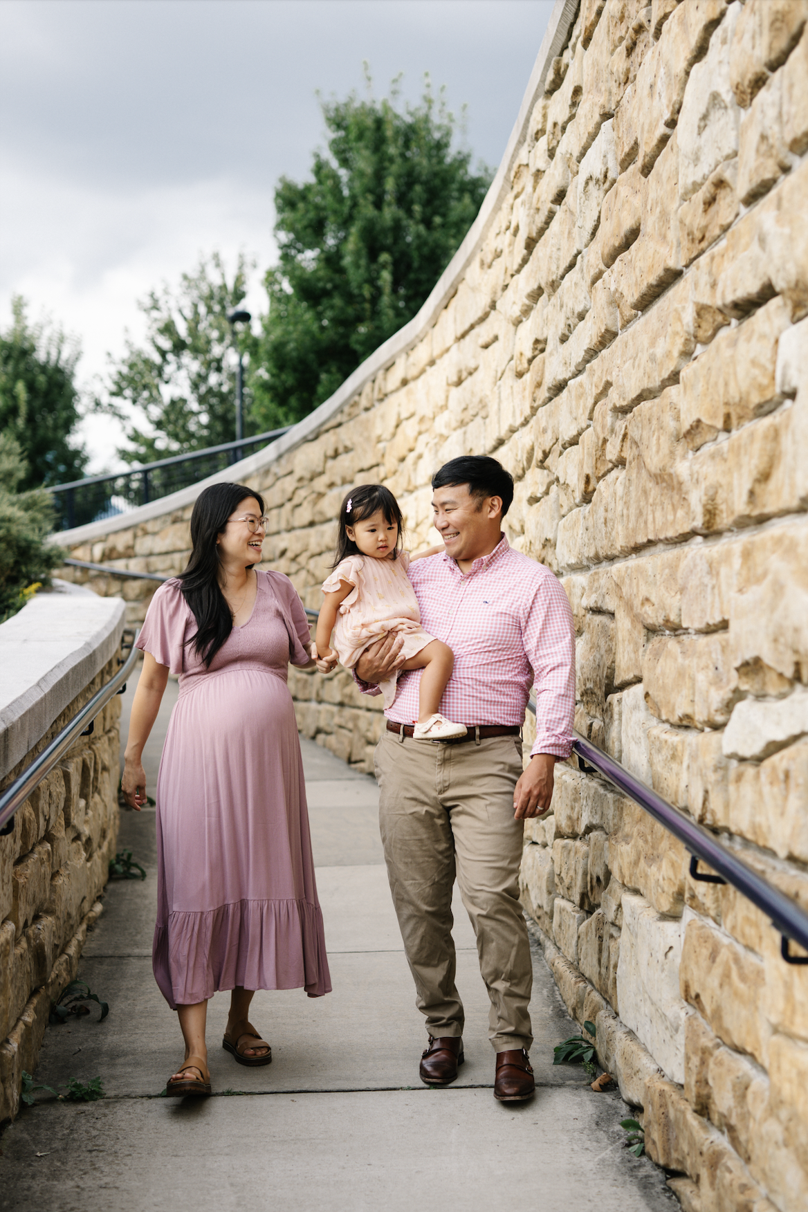 A family of three walking outdoors along a stone wall; the father is carrying a young girl, and the mother is walking beside them, all smiling.