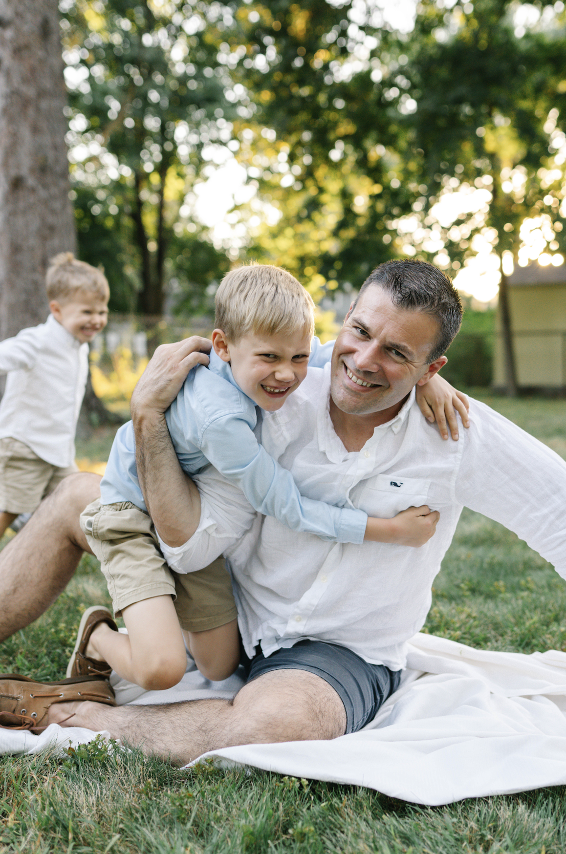 A man and two children playing and laughing outdoors in a grassy backyard with trees in the background during the daytime.