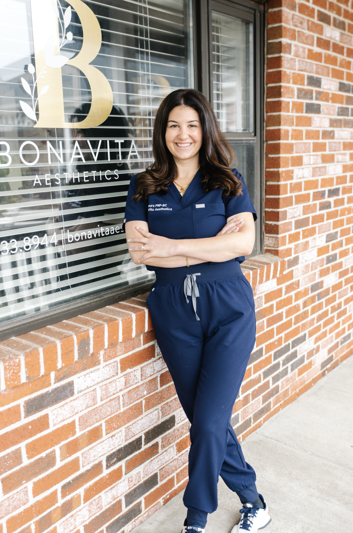 A woman standing outside a brick building, wearing navy scrubs with conference badges, smiling with arms crossed, in front of a window with the Bonavista Aesthetics logo.