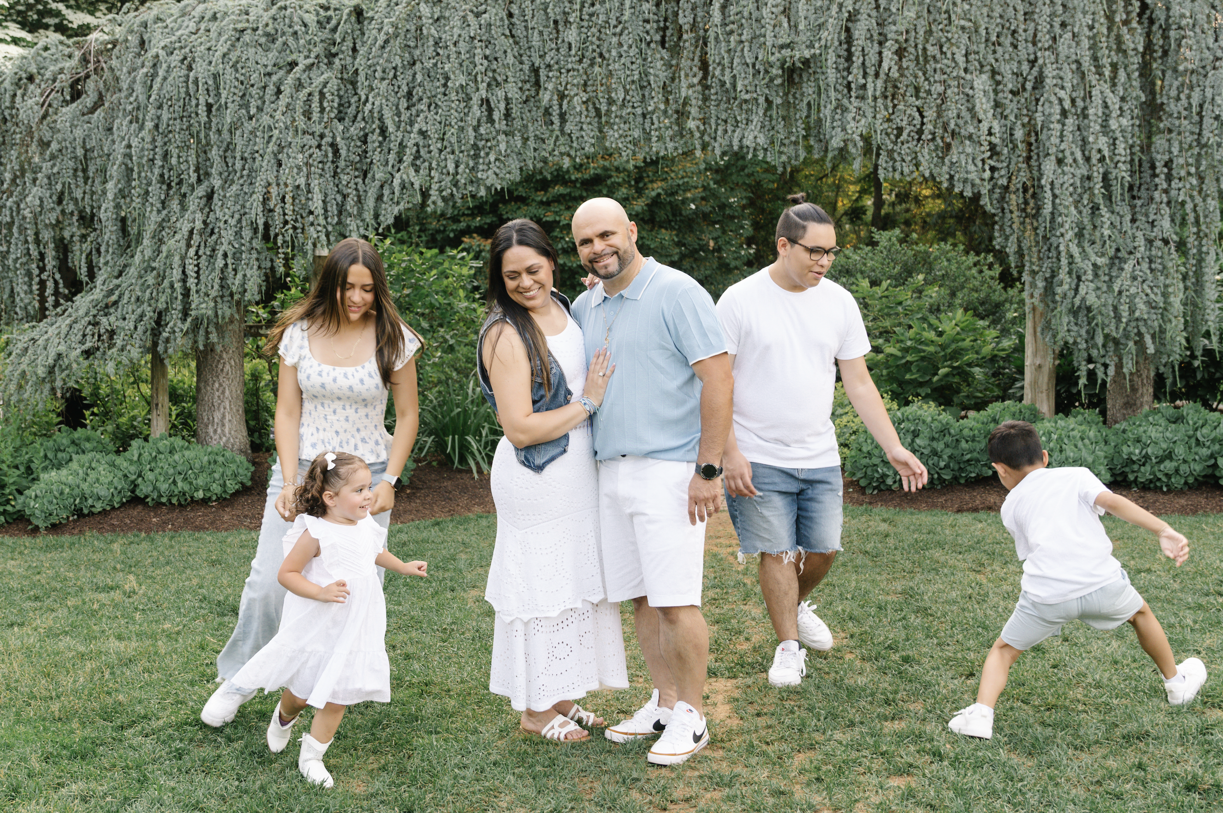 Family of six enjoying time outdoors in a garden with large trees and greenery, some are playing while others are posing for the photo.