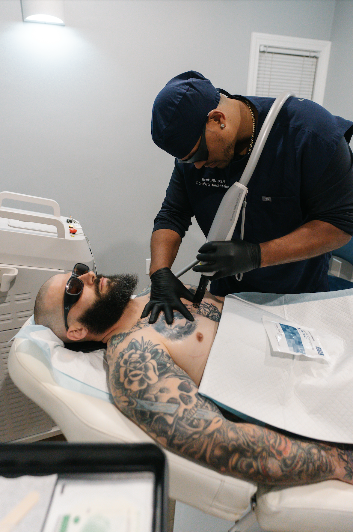 A tattooed man wearing sunglasses lying on a medical bed while a technician performs laser tattoo removal using a handheld laser device on his chest. The technician is wearing black gloves and protective eyewear.