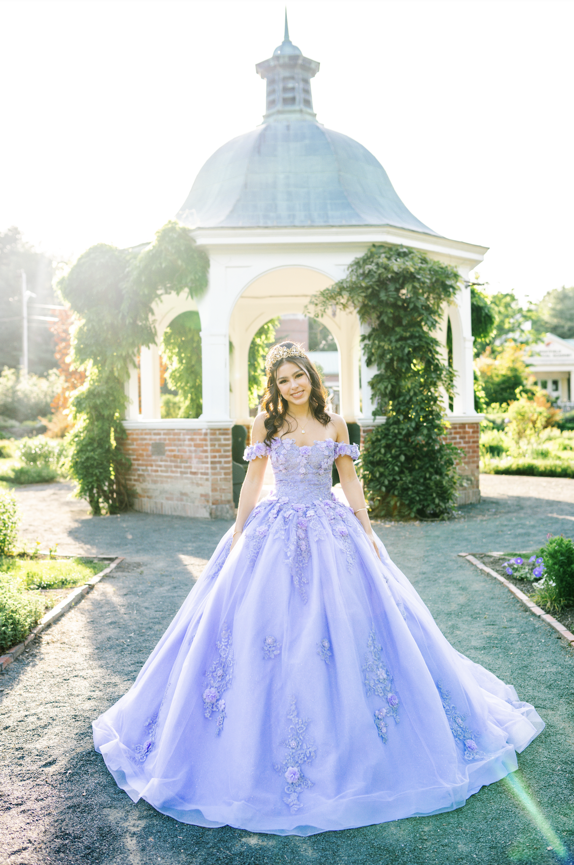 A girl in a lavender gown and tiara stands in front of a white gazebo with brick accents, surrounded by greenery, with sunlight shining from behind.