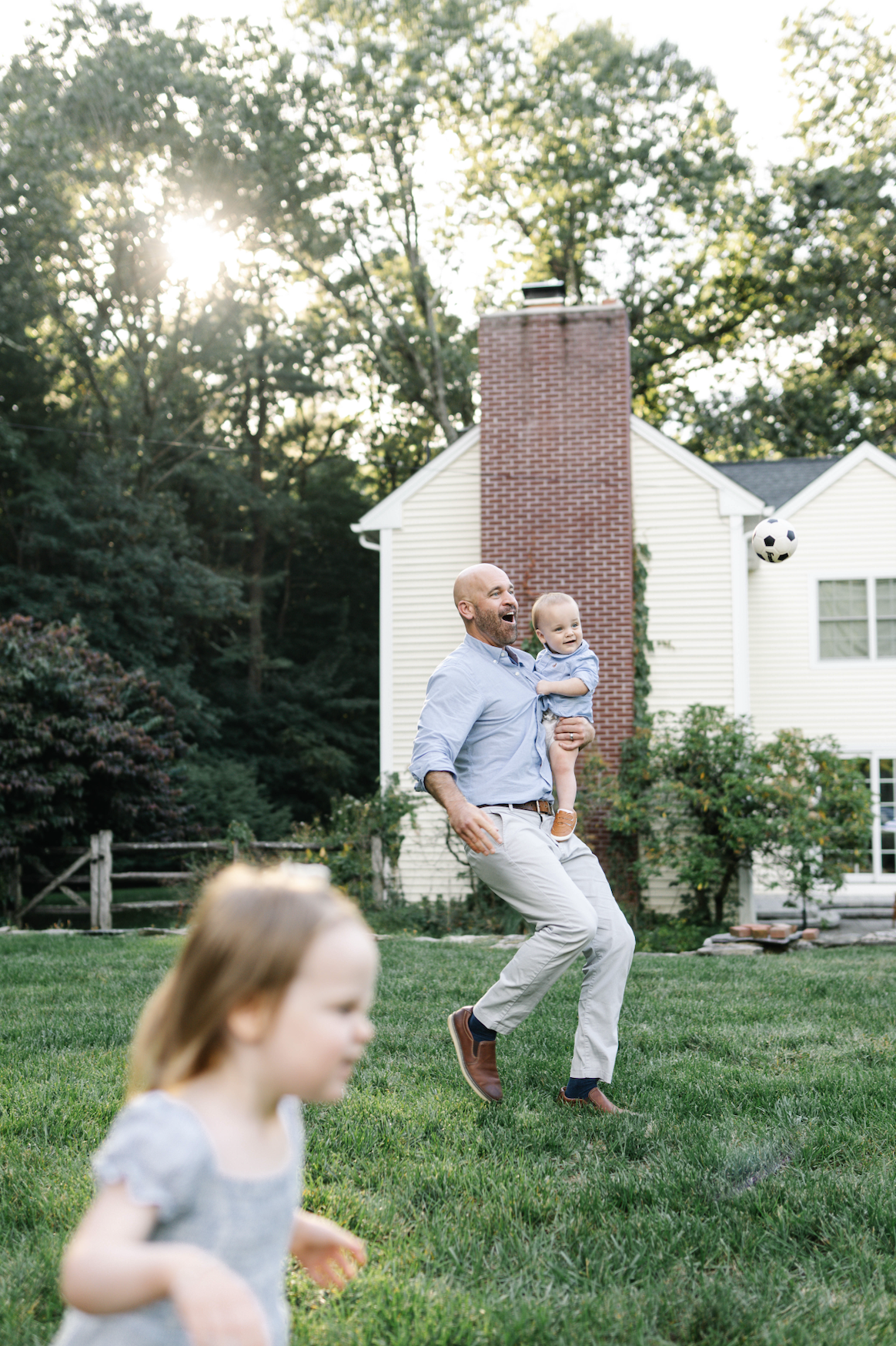 A man with a child in his arms playing with a soccer ball in a backyard, with a girl in the foreground and a house with a chimney in the background.