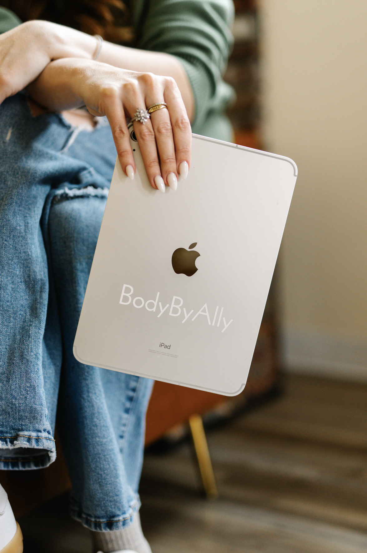 A person with manicured nails and several rings on their fingers holding a white iPad with a logo and the text 'BodyByAlly' on it, sitting with their leg crossed and wearing ripped jeans.