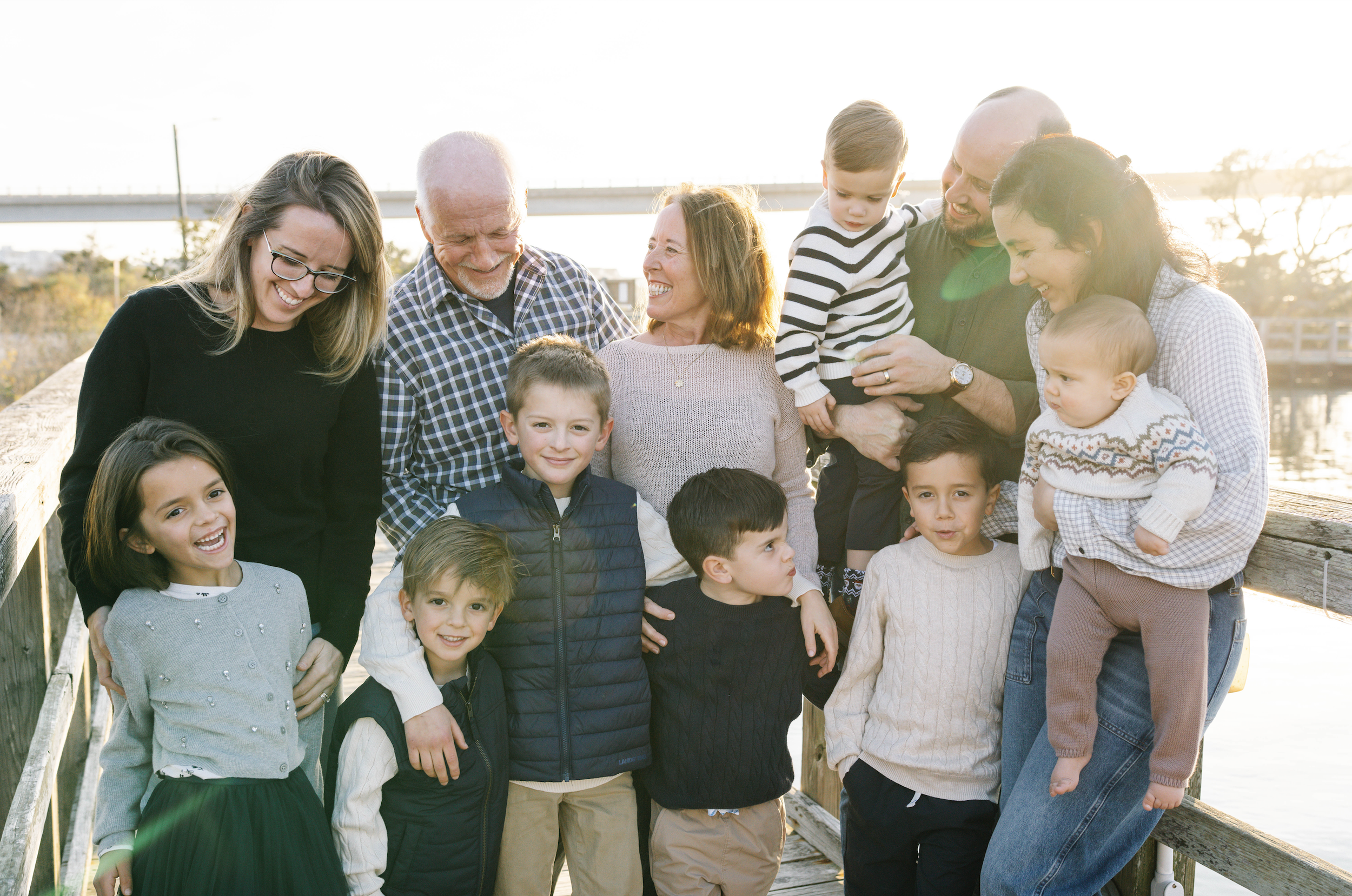 A large multigenerational family of 14 people, including children, teenagers, and adults, gathered on a wooden pier by the water during sunset, smiling and embracing each other.