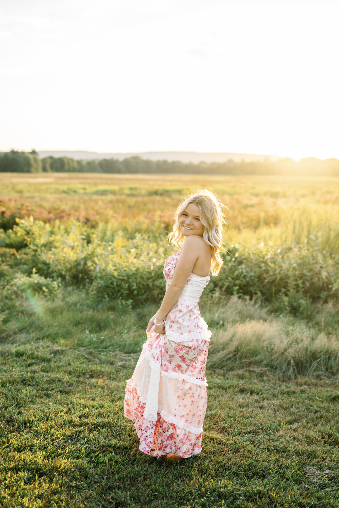 A young woman with blonde curly hair wearing a pink floral dress standing on grass in a field at sunset, smiling at the camera.