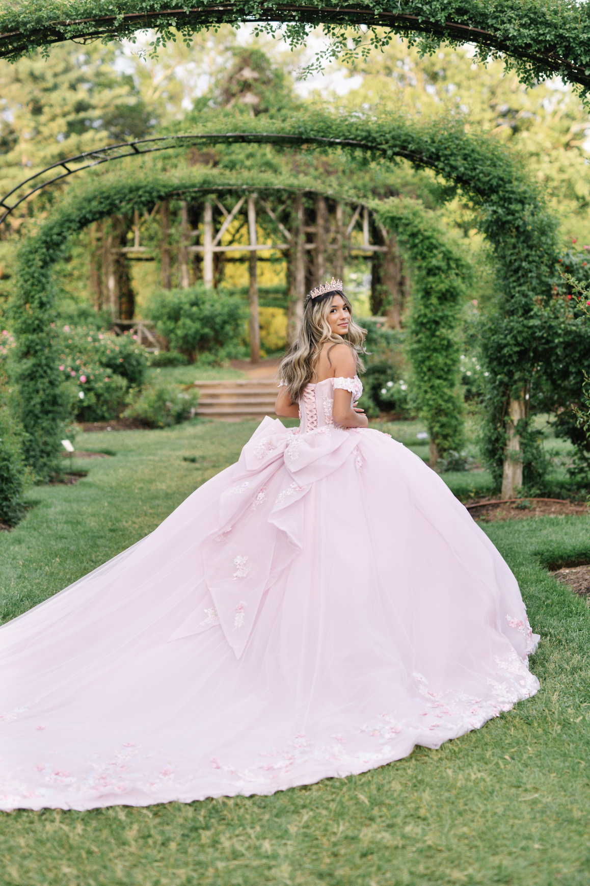 A young woman in a pink princess dress with floral details and a large bow is standing on a grassy area in a garden with green shrubs and trees, under a decorative metal archway covered in greenery, wearing a tiara.