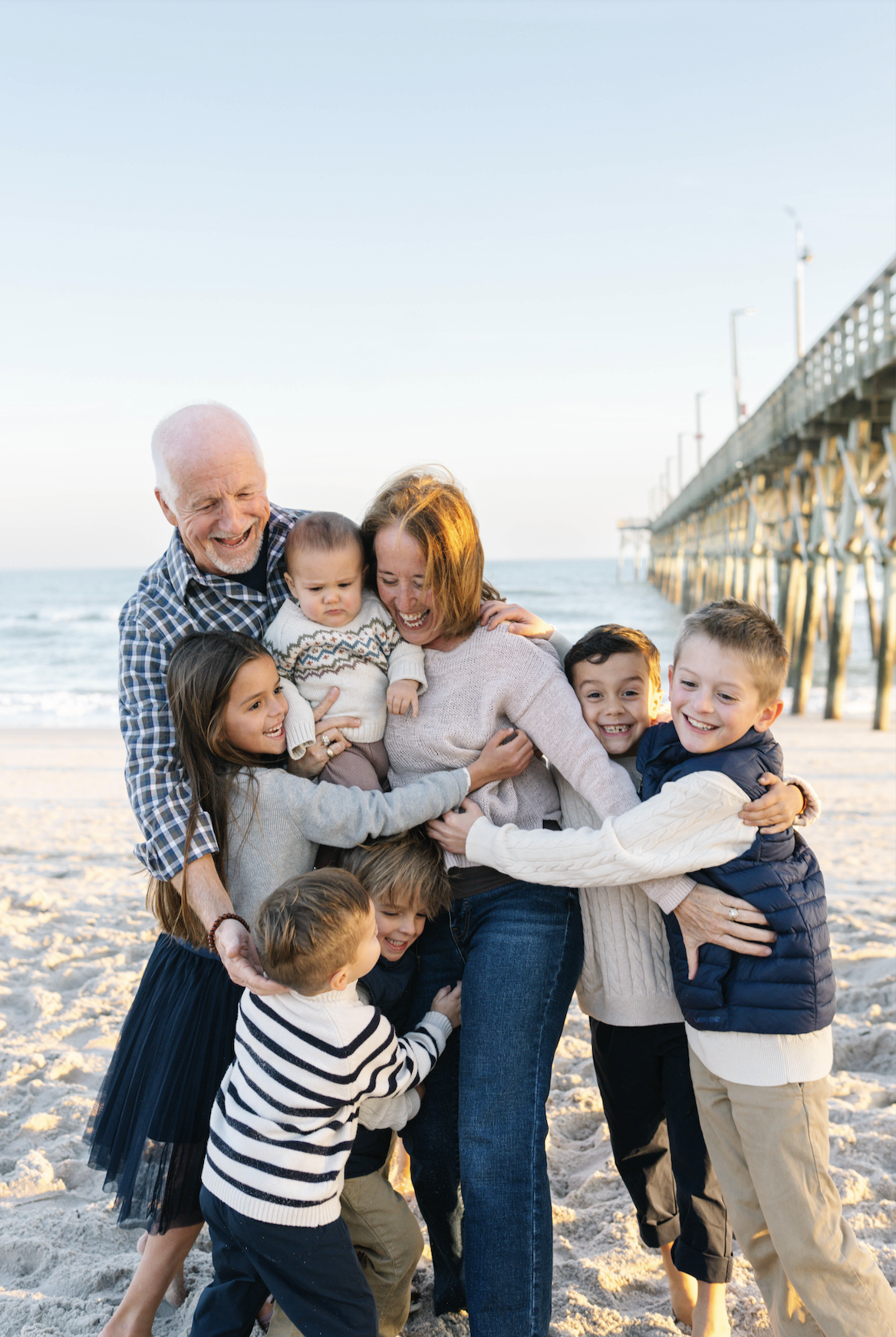 A multigenerational family, including grandparents, parents, and children, hugging and laughing on a beach with a pier in the background during sunset.