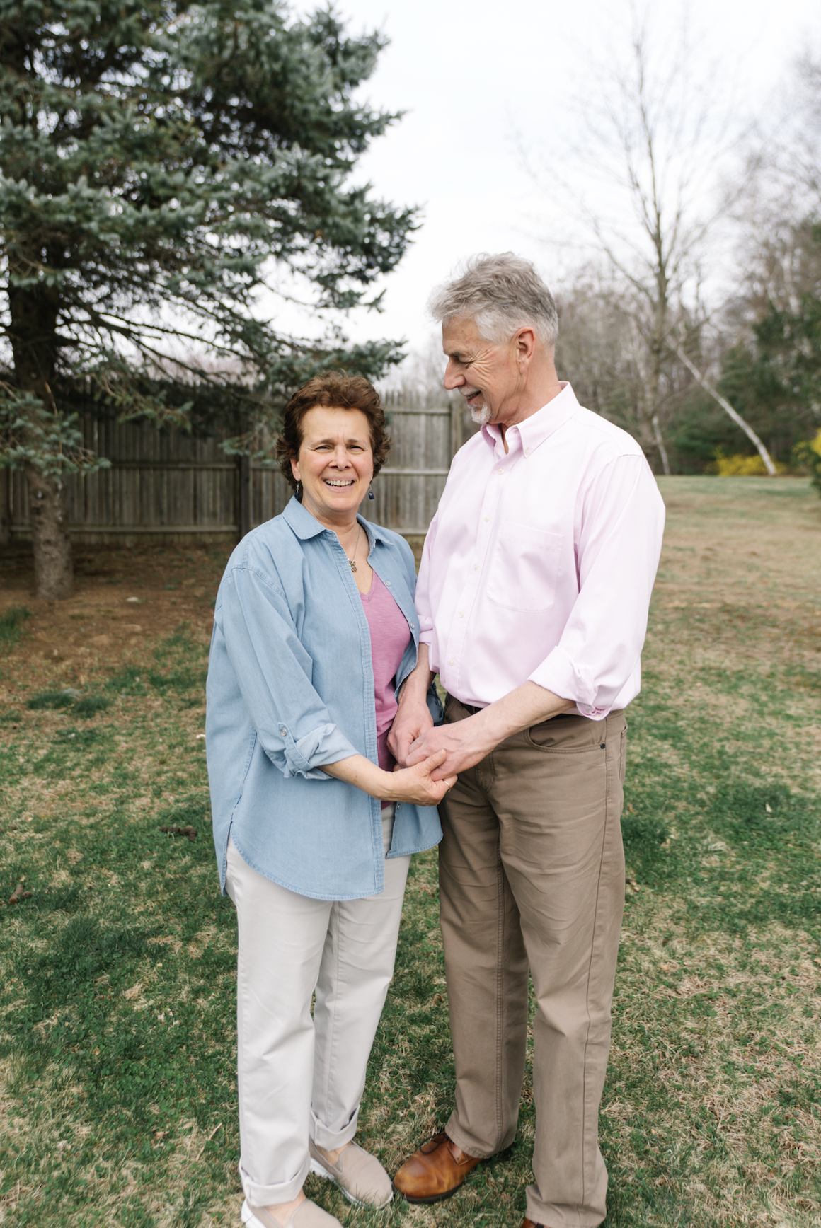An elderly couple holding hands and smiling outdoors on a cloudy day, standing on grass with trees and a wooden fence in the background.