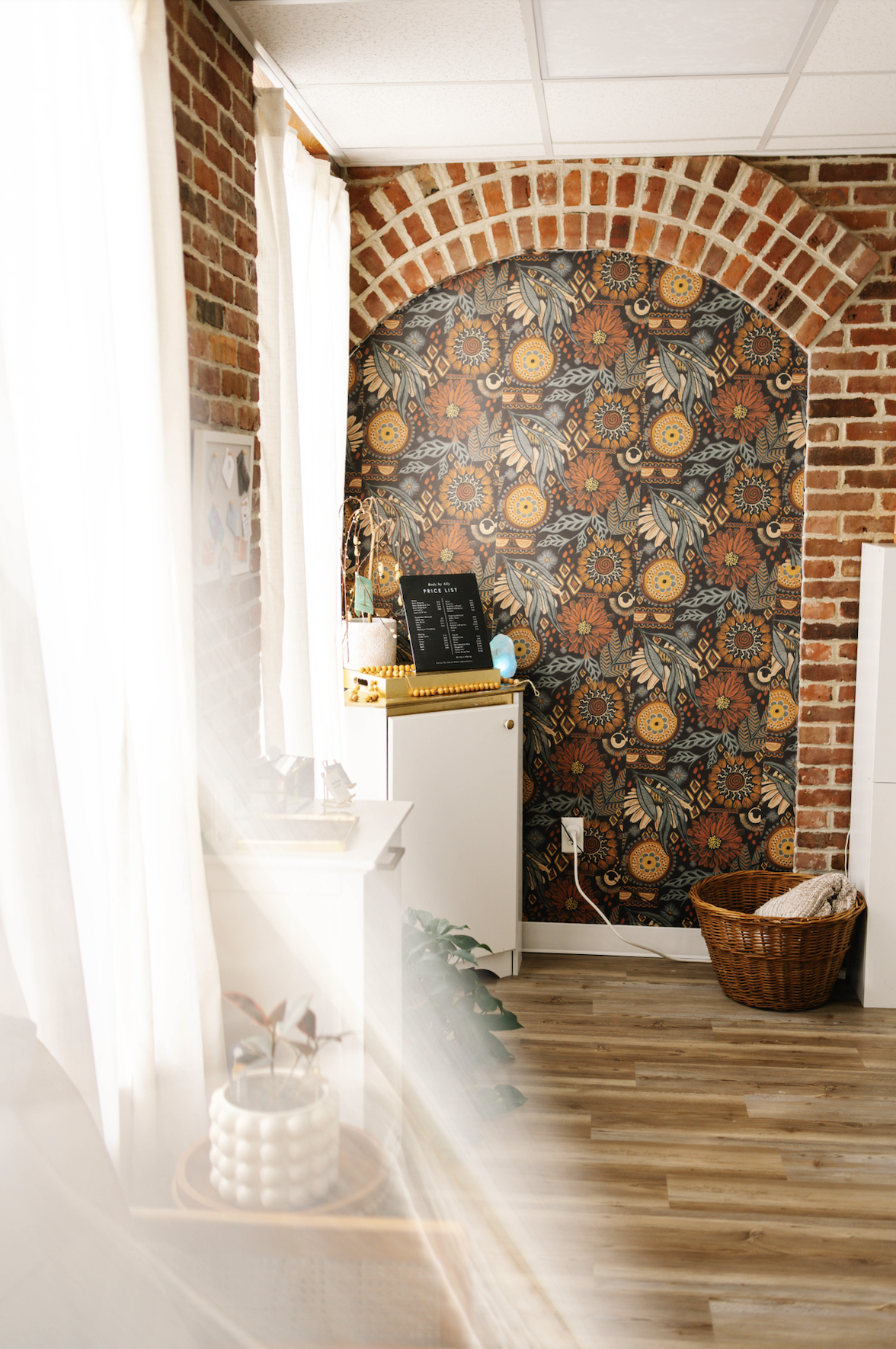 Interior of a room with brick walls, a floral wallpapered accent wall, and white curtains. There is a small white cabinet, a plant in a white textured pot, a basket with cloth, and a price list on a stand.