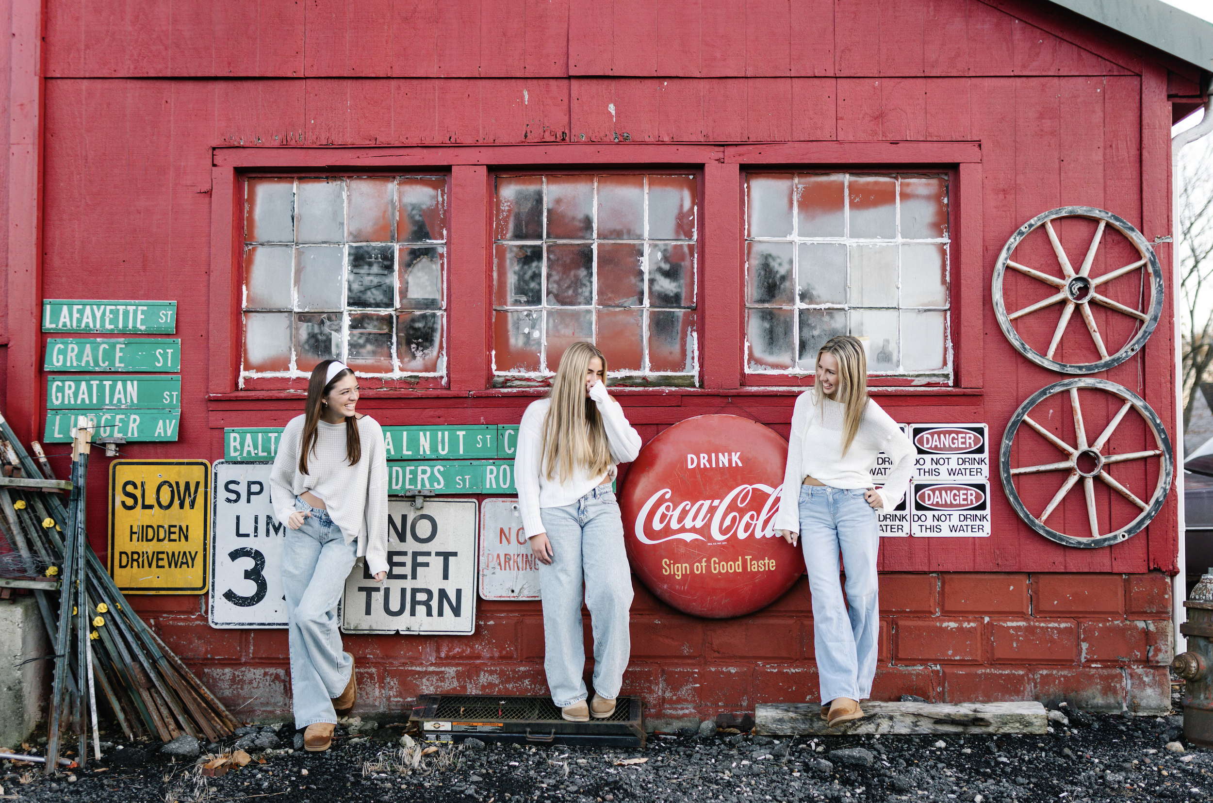 Three women in white sweaters and jeans standing in front of a red barn wall decorated with vintage signs and wheels, with a Coca-Cola sign in the center, and a variety of street name signs on the left.