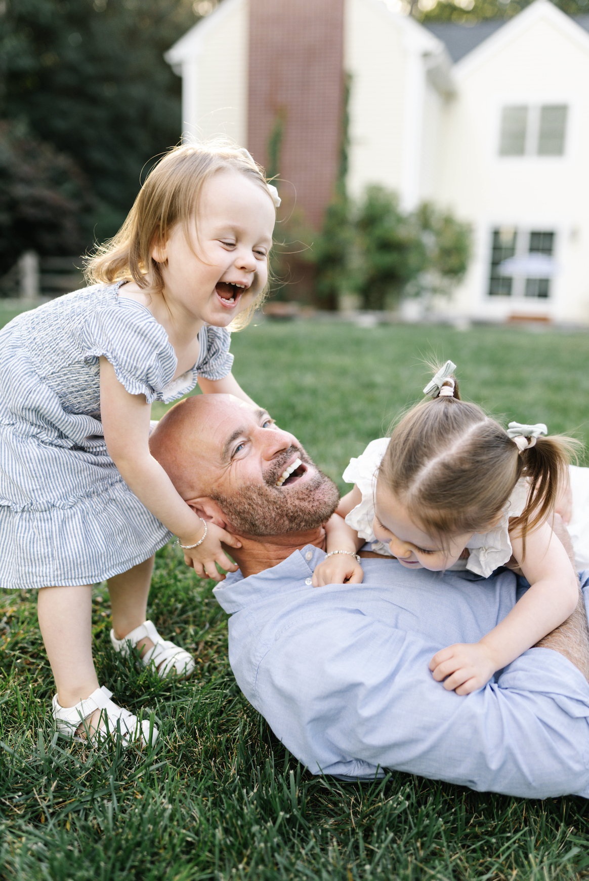 A man lying on the grass playing with two young girls, all smiling and laughing outdoors, in front of a house with a white exterior and large windows.