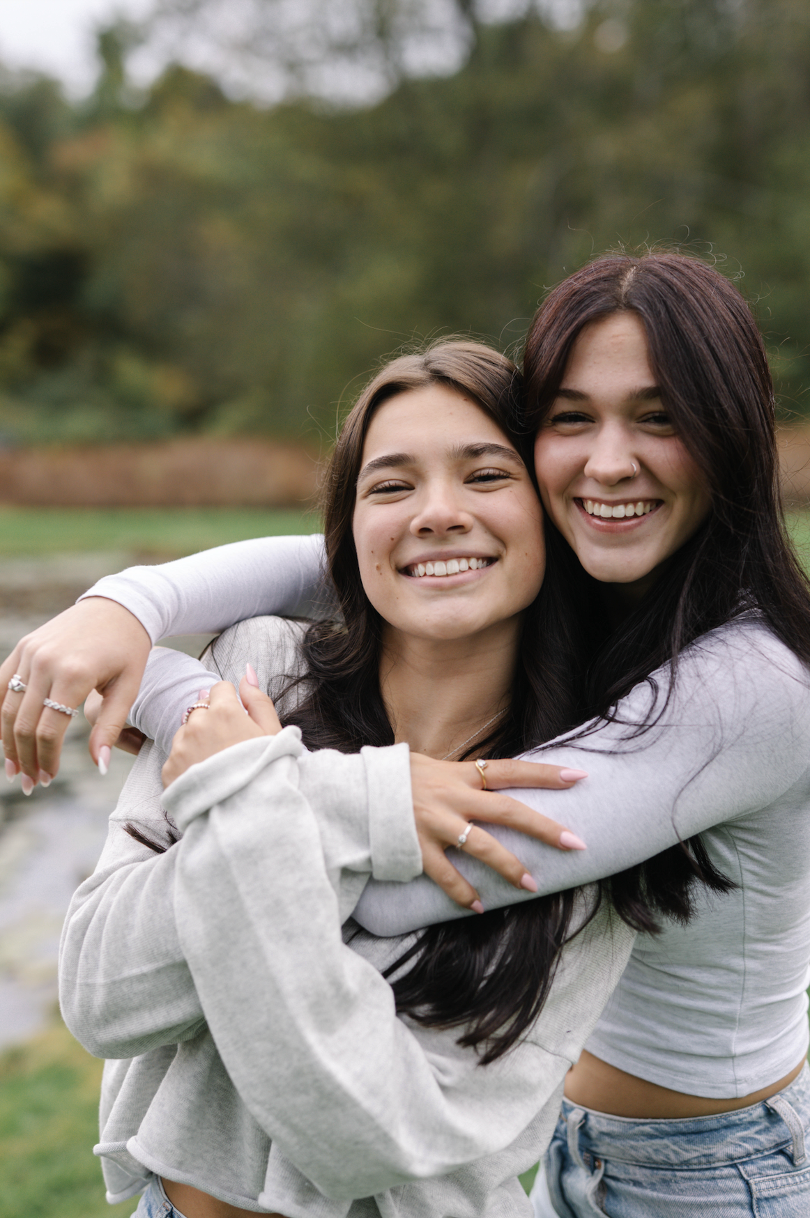 Two young women smiling and hugging outdoors in a park with autumn trees in the background.
