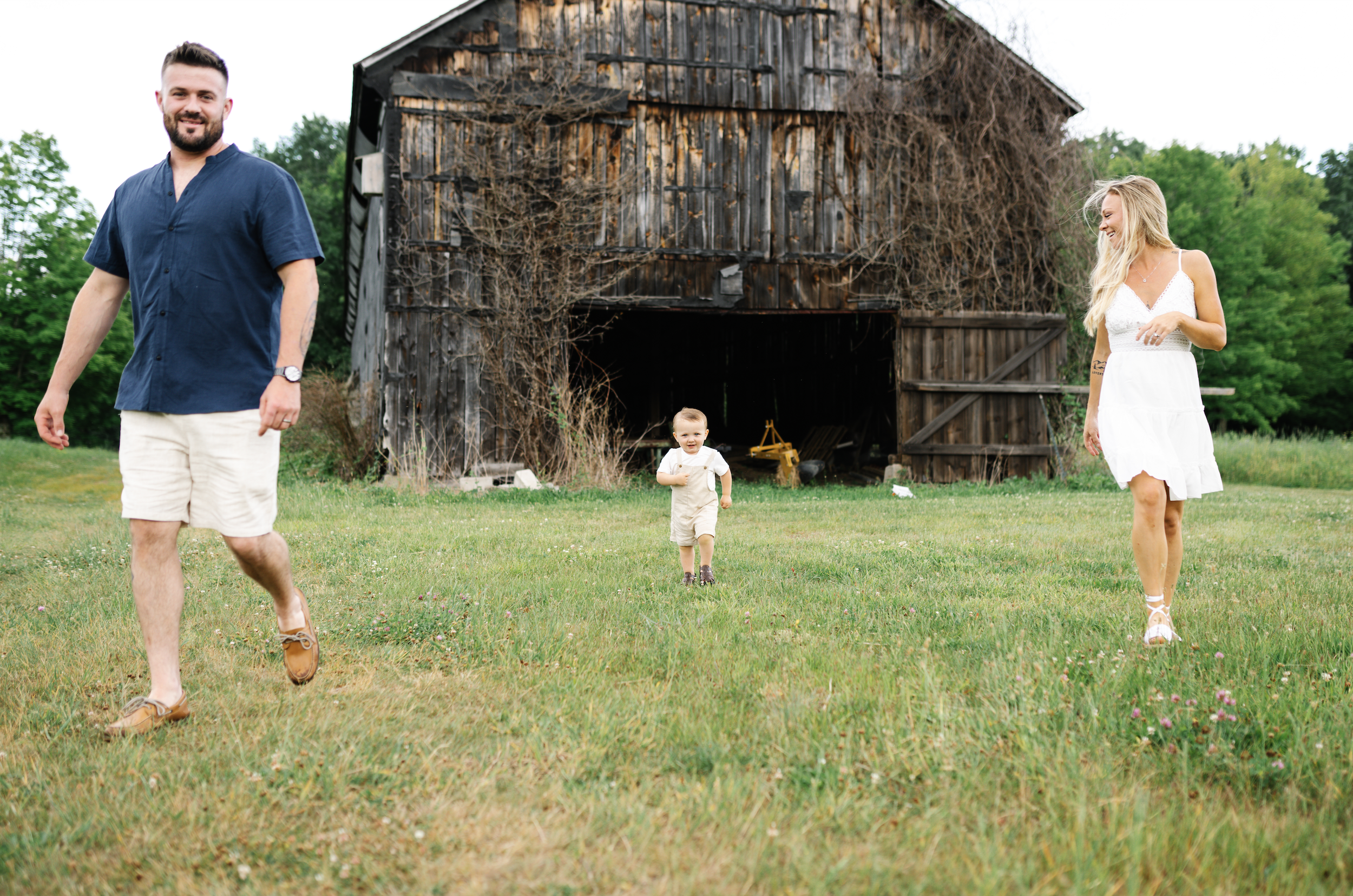 A family of three walking in a grassy field in front of a rustic wooden barn, with green trees in the background. The man is on the left, wearing a navy shirt and khaki shorts, smiling. The woman is on the right, wearing a white dress, smiling and looking at the young child in the center, who is running towards the camera, dressed in a white shirt and beige overalls.