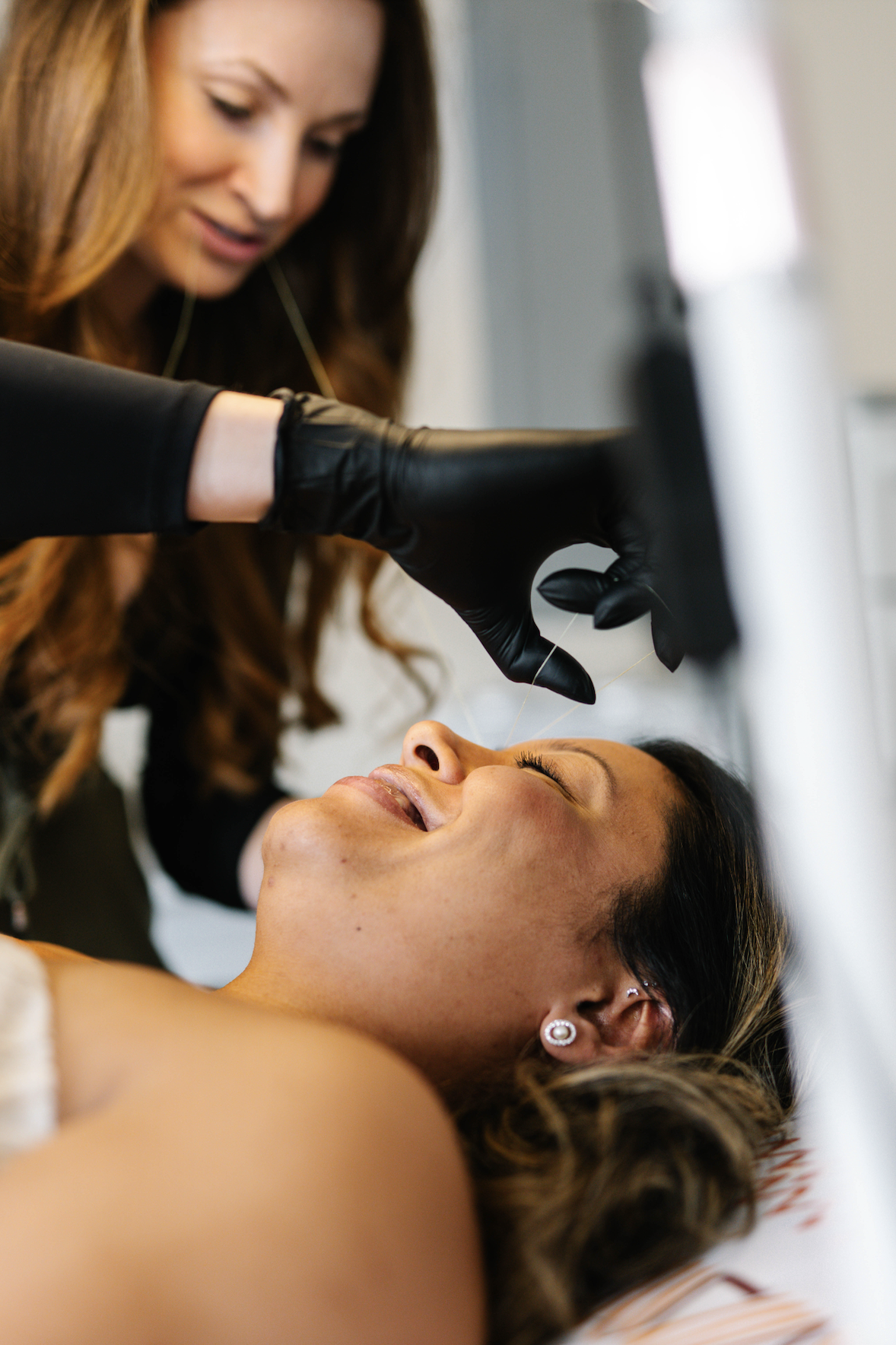 A woman lying down with her eyes closed and a smile, receiving a facial from another woman wearing black gloves, in a spa or salon setting.