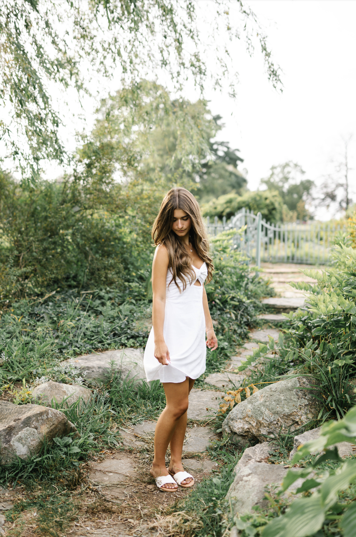A young woman in a white dress and sandals walks on a garden path surrounded by greenery.