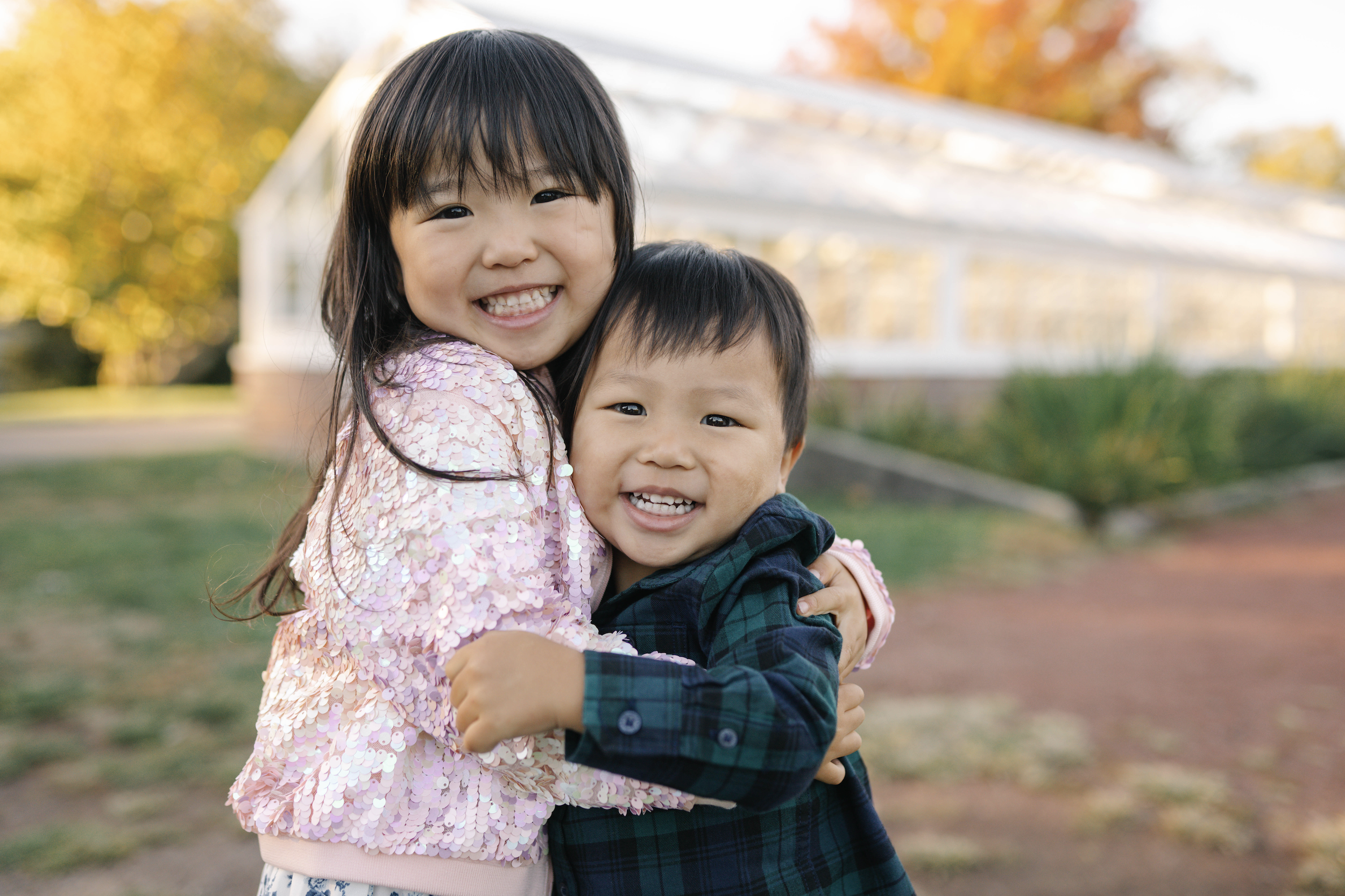 A young girl and a young boy happily hugging outdoors with trees and a white building in the background.