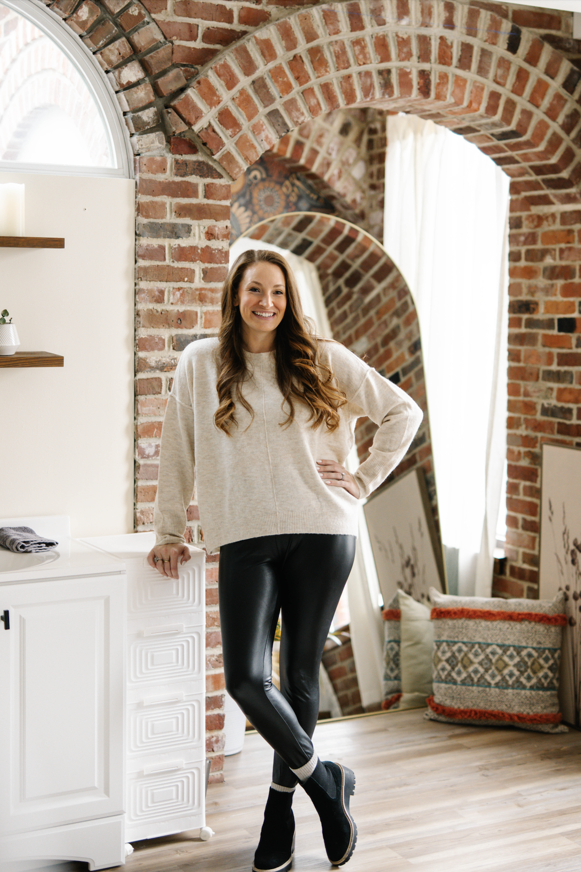 A smiling woman stands in a cozy room with exposed brick walls and arched ceilings, wearing a cream sweater, black shiny leggings, and slip-on shoes.