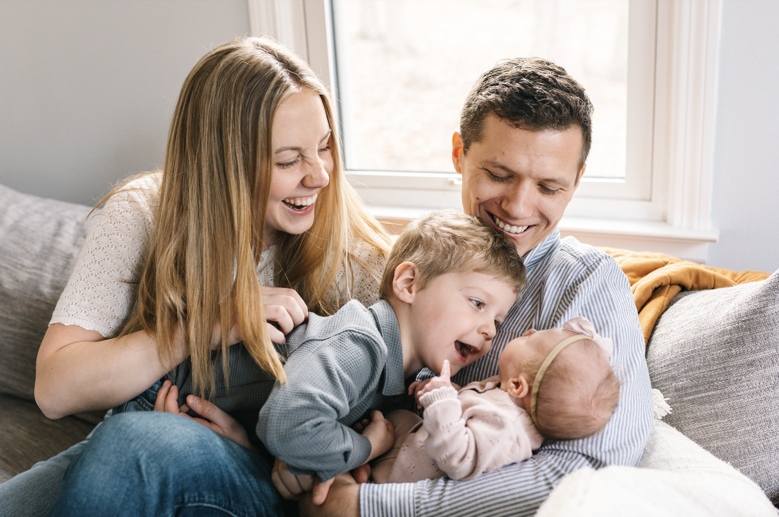 A family of four with a mother, father, young boy, and baby girl sitting on a couch near a window, smiling and playing together.