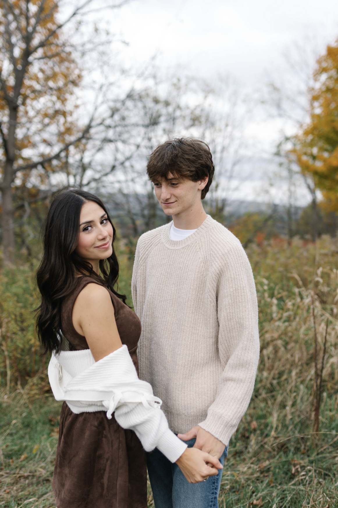 A young couple standing outdoors in an autumn setting, holding hands and looking at the camera, with fall foliage in the background.