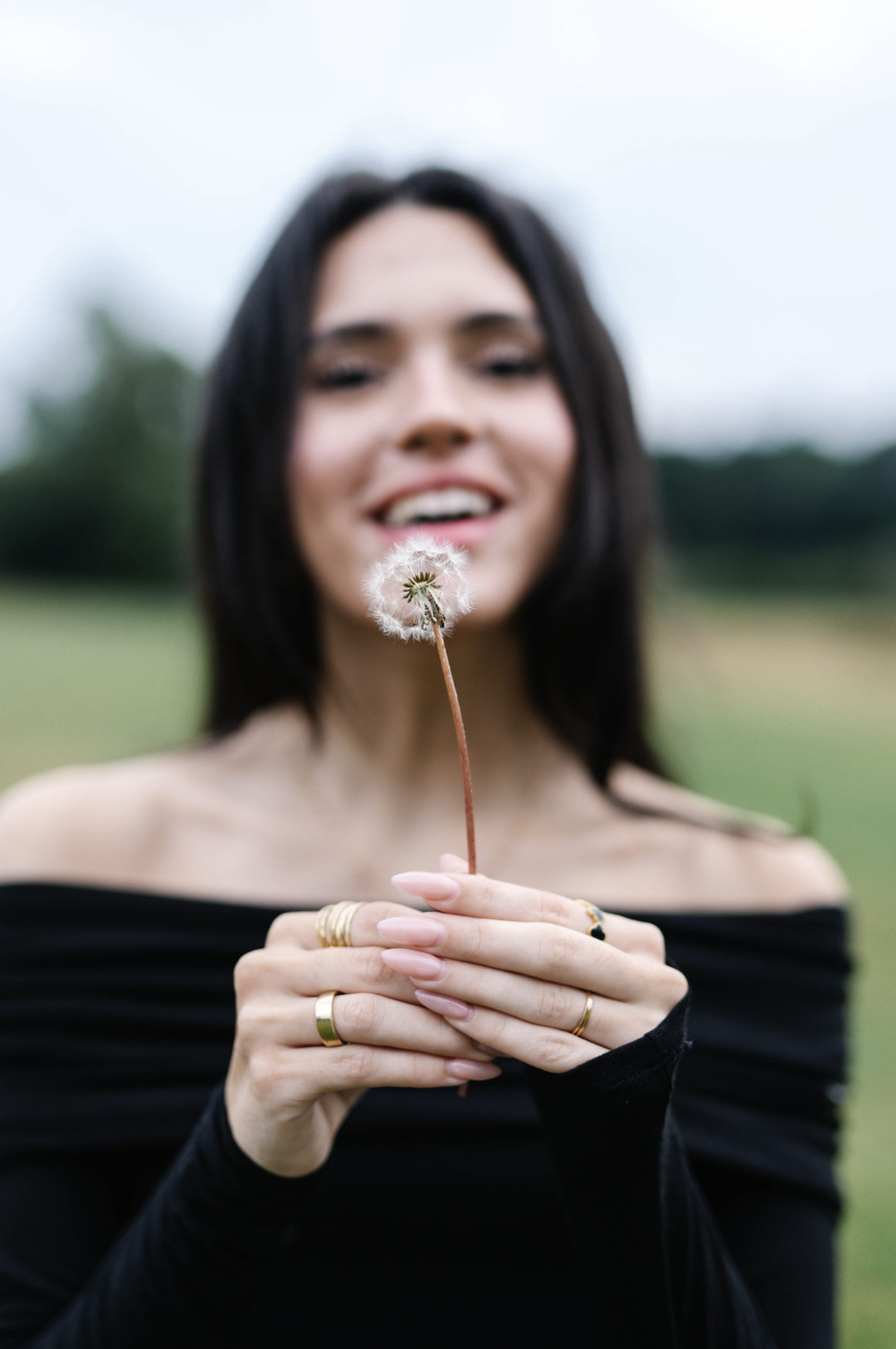 Woman with long dark hair holding a dandelion flower in an open field, smiling