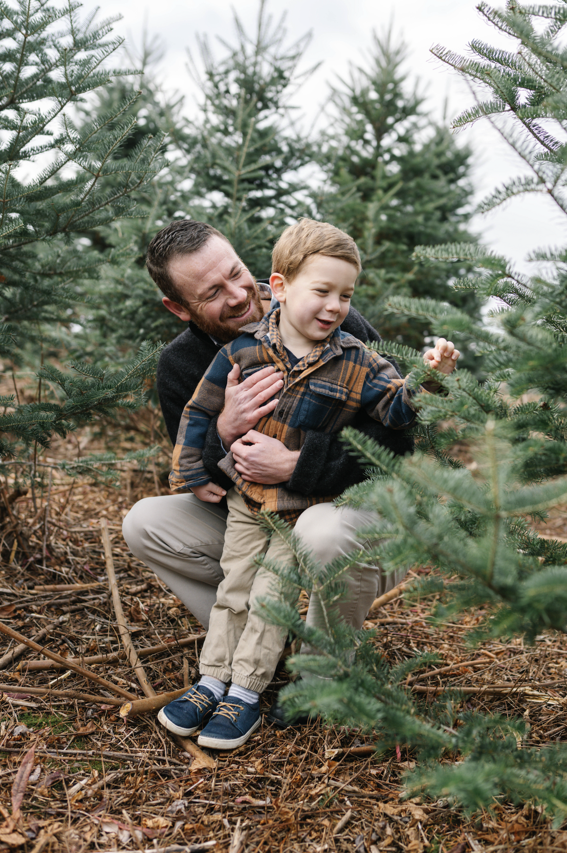 A man and young boy enjoy a walk among Christmas trees at a tree farm, with fall leaves on the ground and overcast sky.
