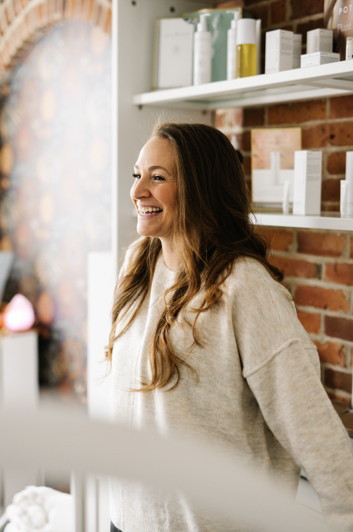 A smiling woman with long brown hair in a beige sweater standing indoors near a brick wall and shelves with skincare products.
