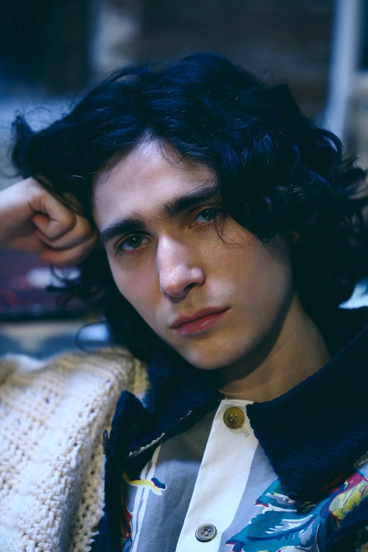 Close-up portrait of a person with shoulder-length wavy dark hair, light skin, and blue eyes, looking directly at the camera with a contemplative expression, resting their head on their hand.