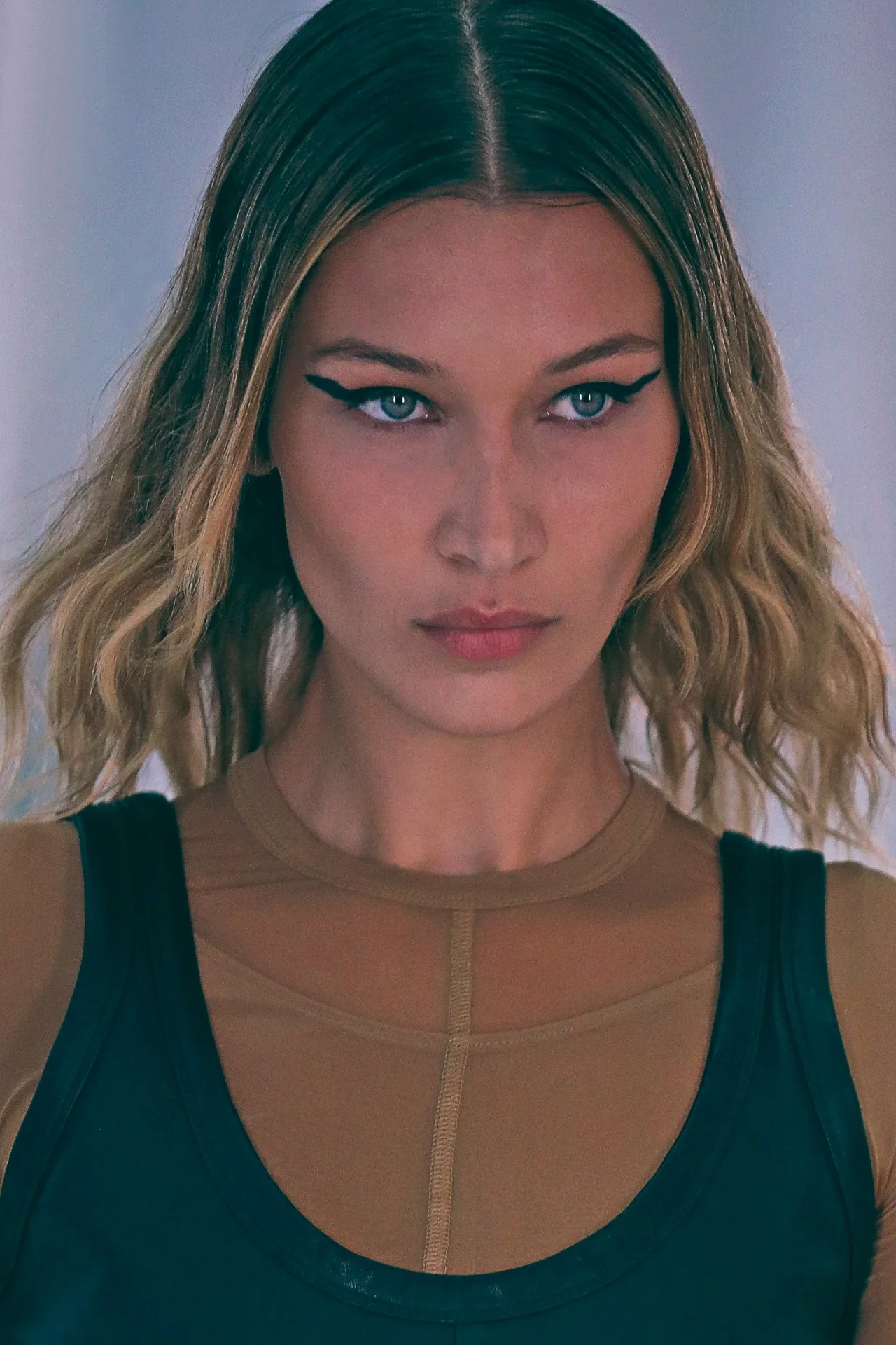 A woman with wavy blonde hair, blue eyes, and winged eyeliner looking directly at the camera, wearing a black top with a beige or nude-colored top underneath.