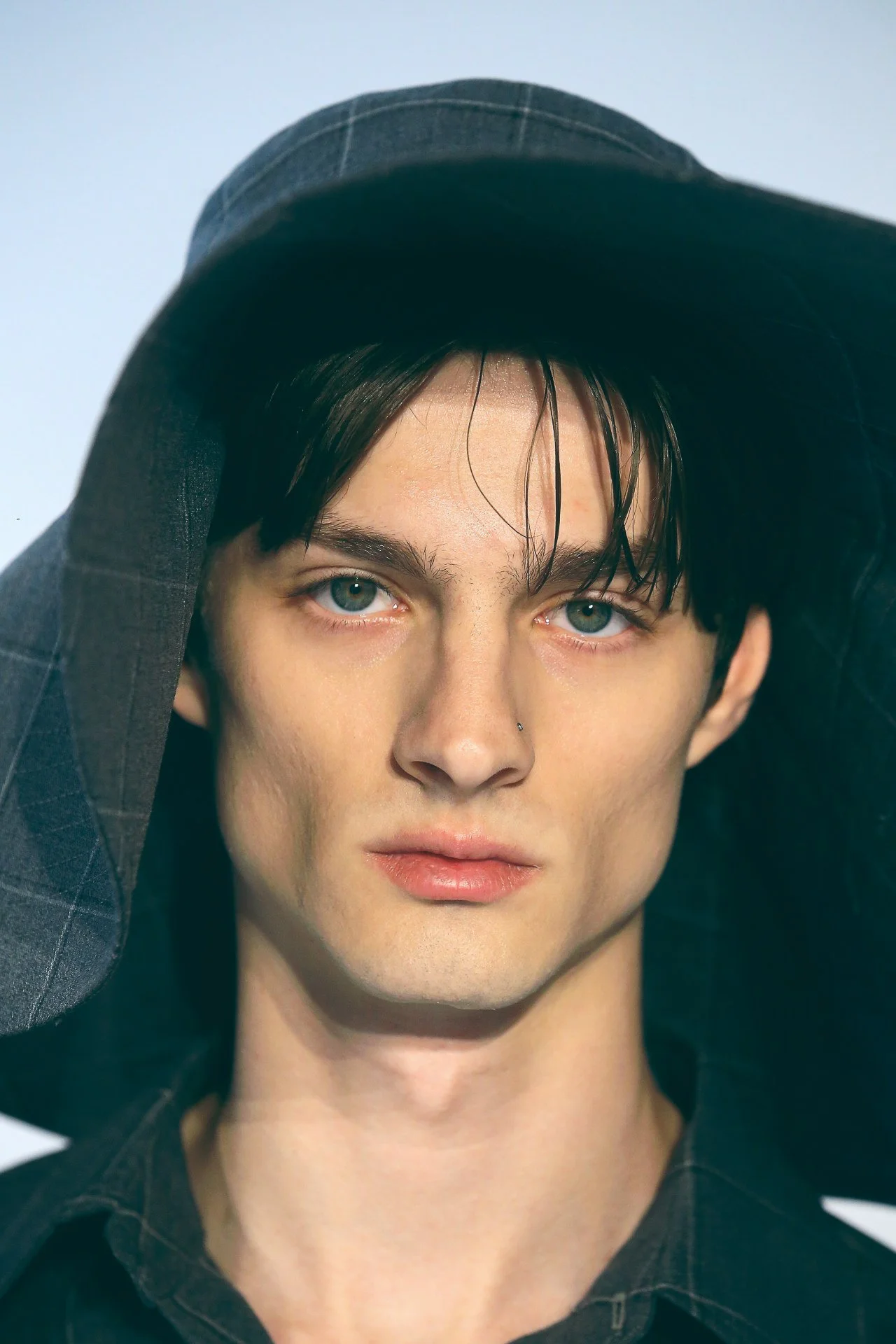 A person with short dark hair, piercing blue eyes, and a nose piercing, wearing a dark-colored shirt and a checkered hat, holding the brim of the hat with one hand.