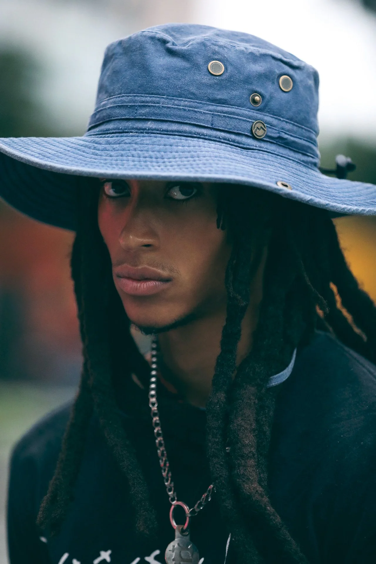 A person with dreadlocks wearing a large blue wide-brimmed hat with metal pins and a black shirt, looking at the camera with a serious expression.