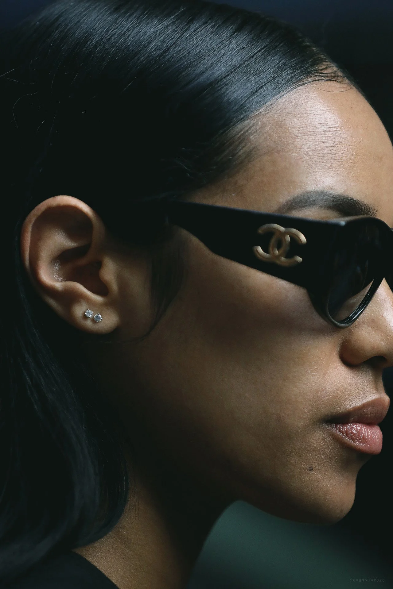 Close-up side profile of a woman wearing black Chanel sunglasses, diamond stud earrings, with sleek dark hair, and smooth skin.