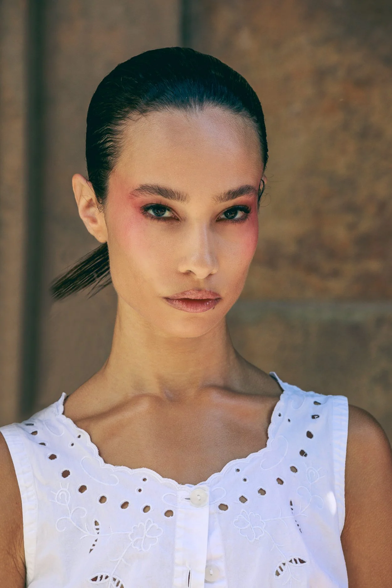 A woman with dark, wet-look hair slicked back, wearing a white sleeveless top with eyelet embroidery, standing against a textured brown background.