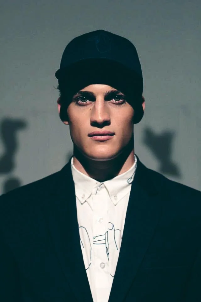 Portrait of a young man with dark eye makeup, wearing a black cap, white button-up shirt with a simple line drawing, and a black blazer, against a plain background.