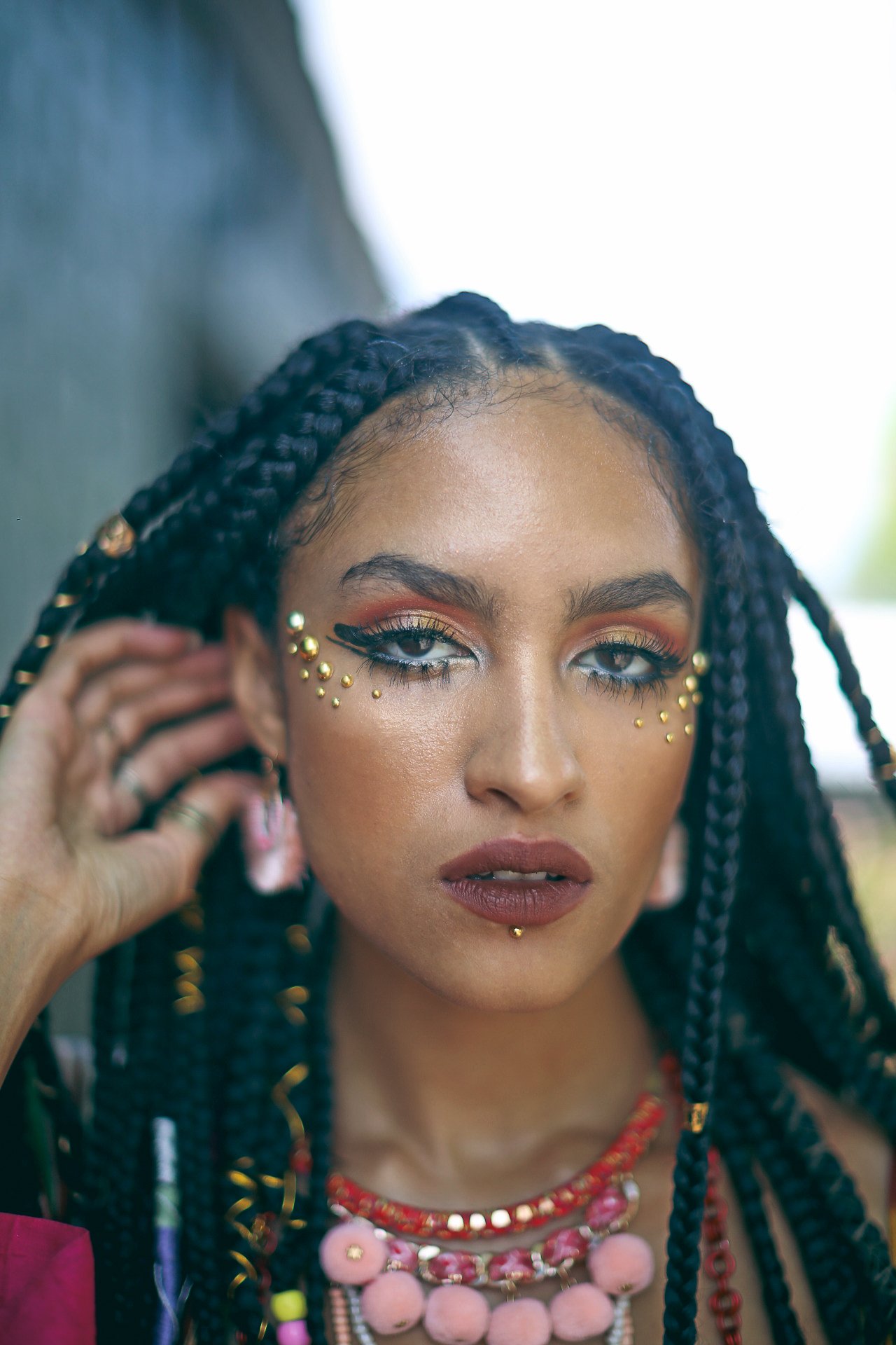 A woman with braided hair and makeup featuring gold embellishments and colorful eyeshadow, wearing layered necklaces and earrings.