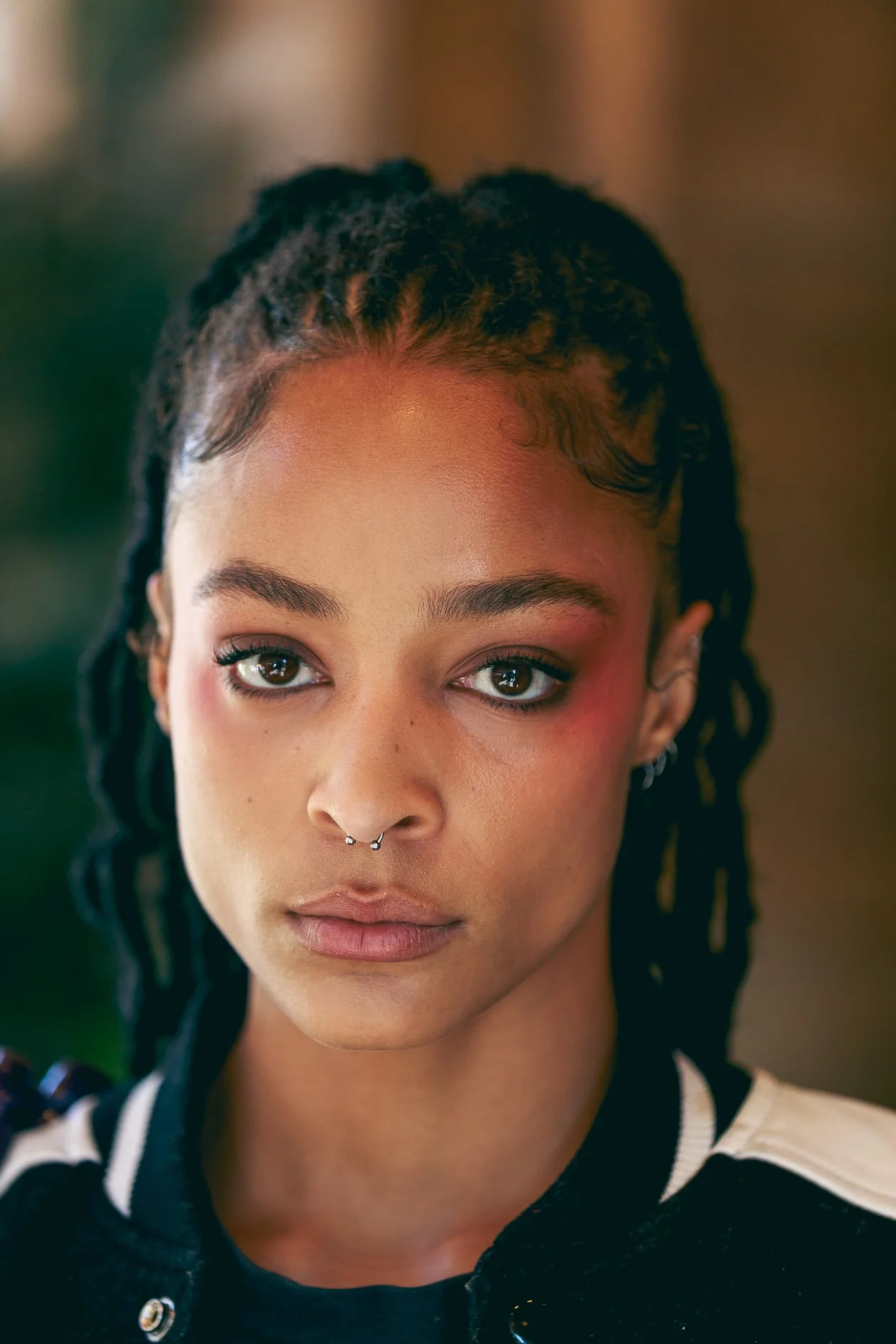 Close-up of a young woman with dreadlocks, wearing a nose ring, with natural makeup and a serious expression, wearing a black and white jacket.