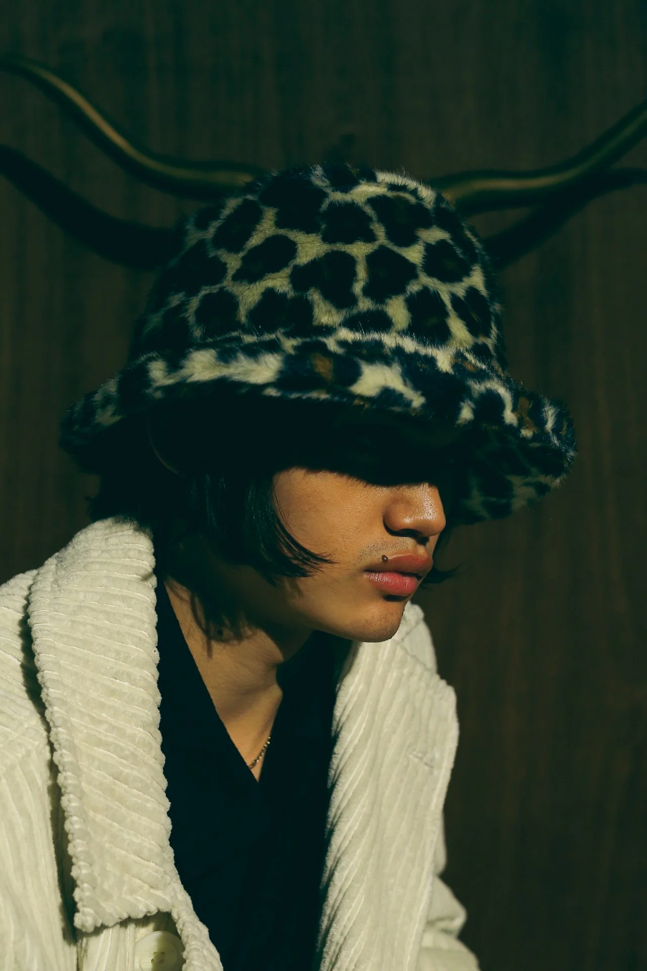 A young person with shoulder-length black hair wearing a leopard print fuzzy hat and a white textured coat, sitting against a dark wooden background with a deer antler mounted on the wall.