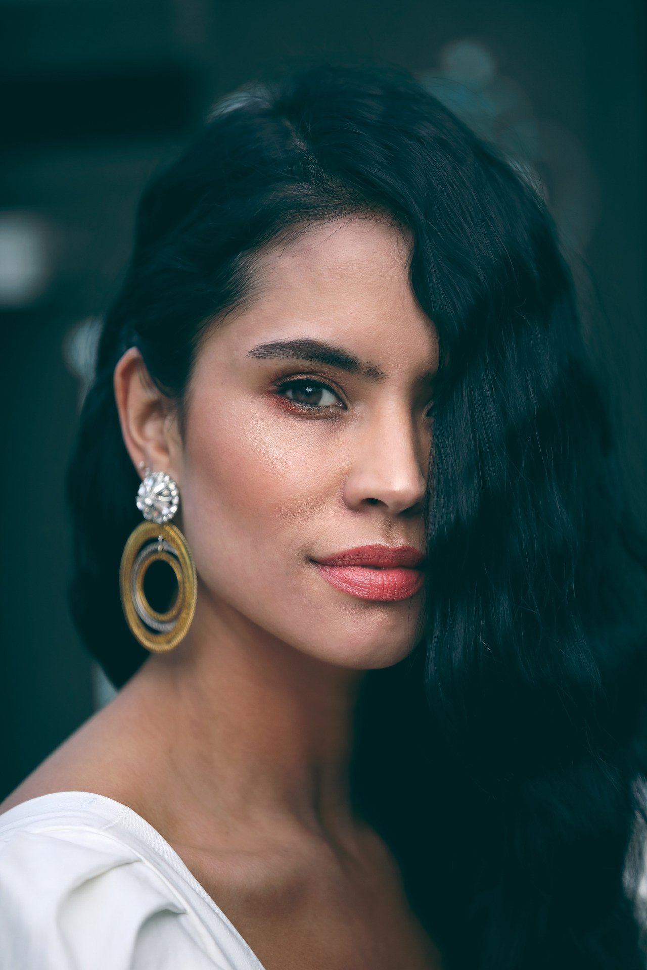 Close-up portrait of a woman with dark, wavy hair partially covering her face, wearing large earrings and a white top, with a confident expression.