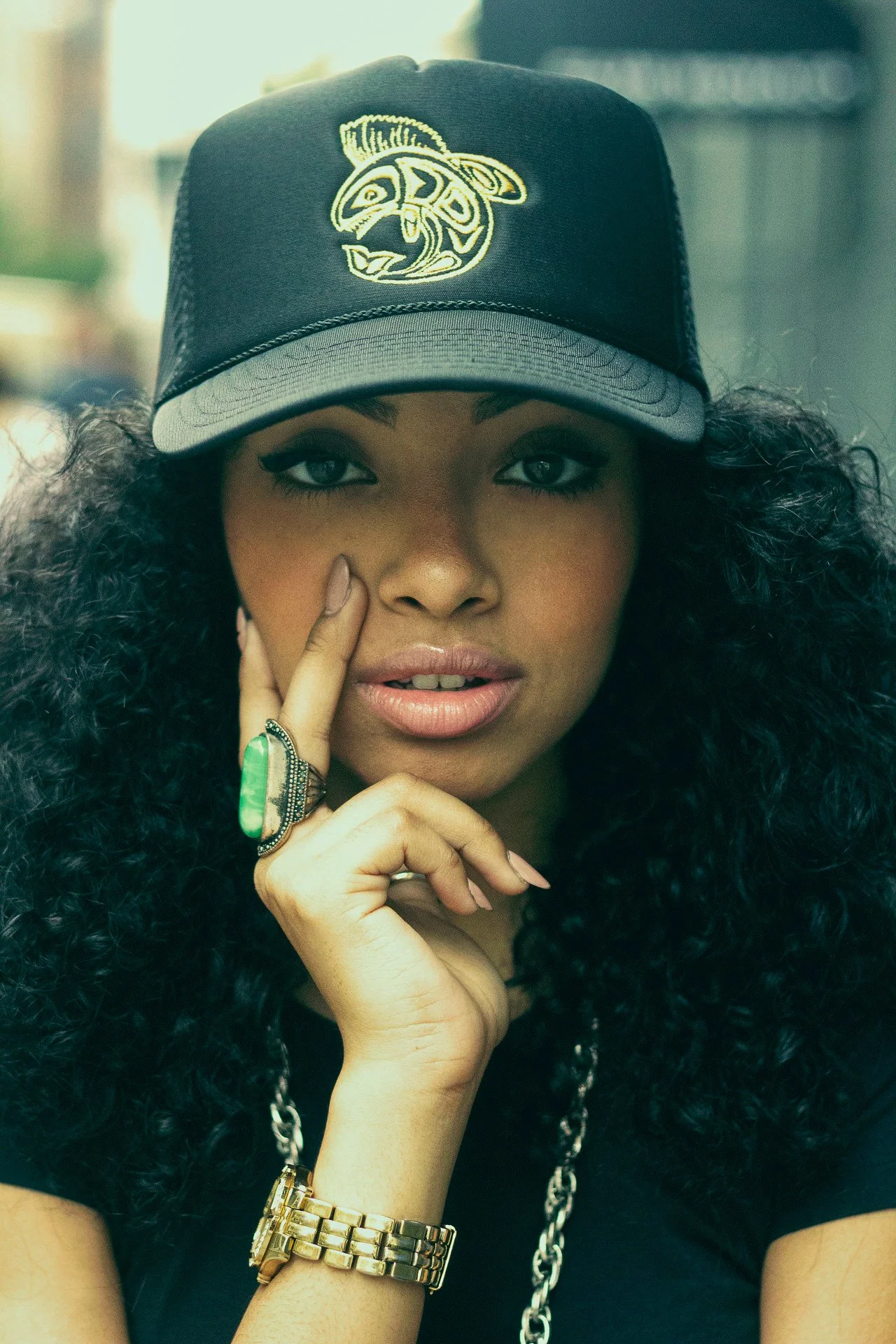 Close-up of a woman with curly black hair wearing a black baseball cap with a yellow and white logo, jewelry including a large ring with a green stone, a gold wristwatch, and a chain necklace, touching her face with her left hand.
