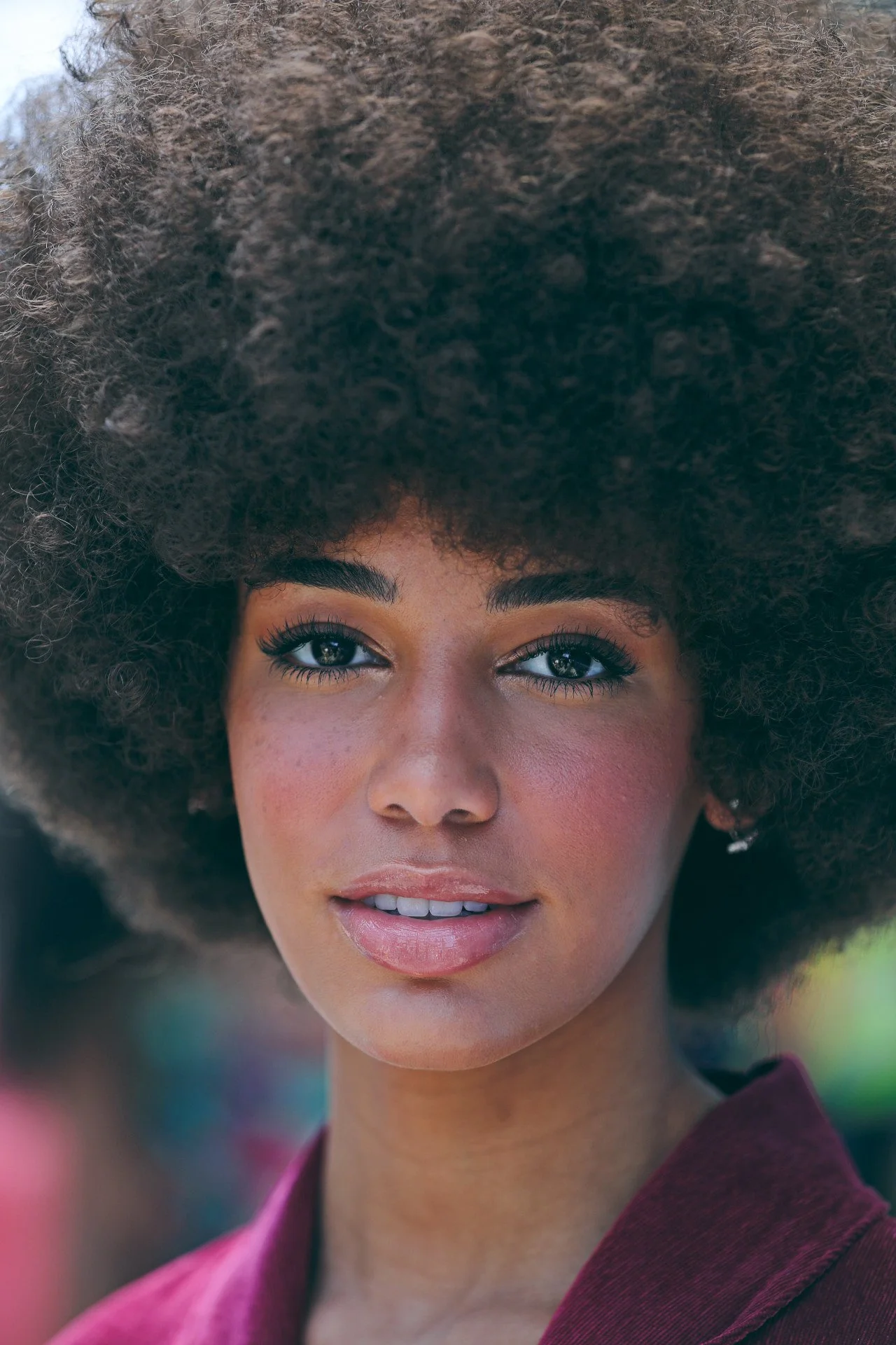 Close-up of a woman with dark brown skin and a large, curly afro hairstyle, wearing a maroon blazer and earrings, looking at the camera with a slight smile.